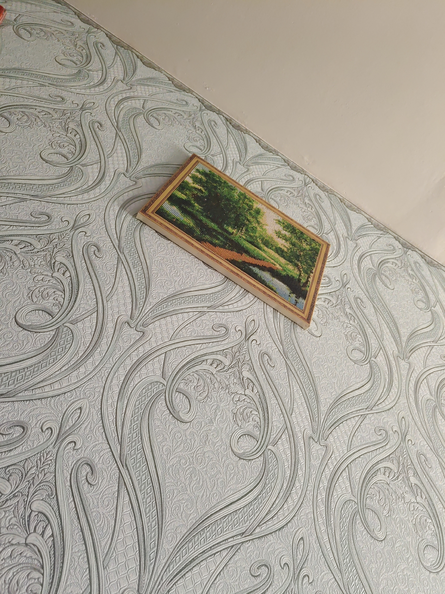 A framed, rectangular art piece, possibly a diamond painting, lies on a wall covered in textured, patterned wallpaper. The art piece depicts a landscape scene with trees and a body of water, illuminated by a soft light, suggesting a natural setting. The wallpaper has a swirling, ornate pattern in a light grey or blue hue. The scene appears to be indoors, with a portion of a plain, light-colored ceiling visible in the upper right corner. The image does not provide specific details that would allow for the identification of a location within the city of Курашим, Russia.