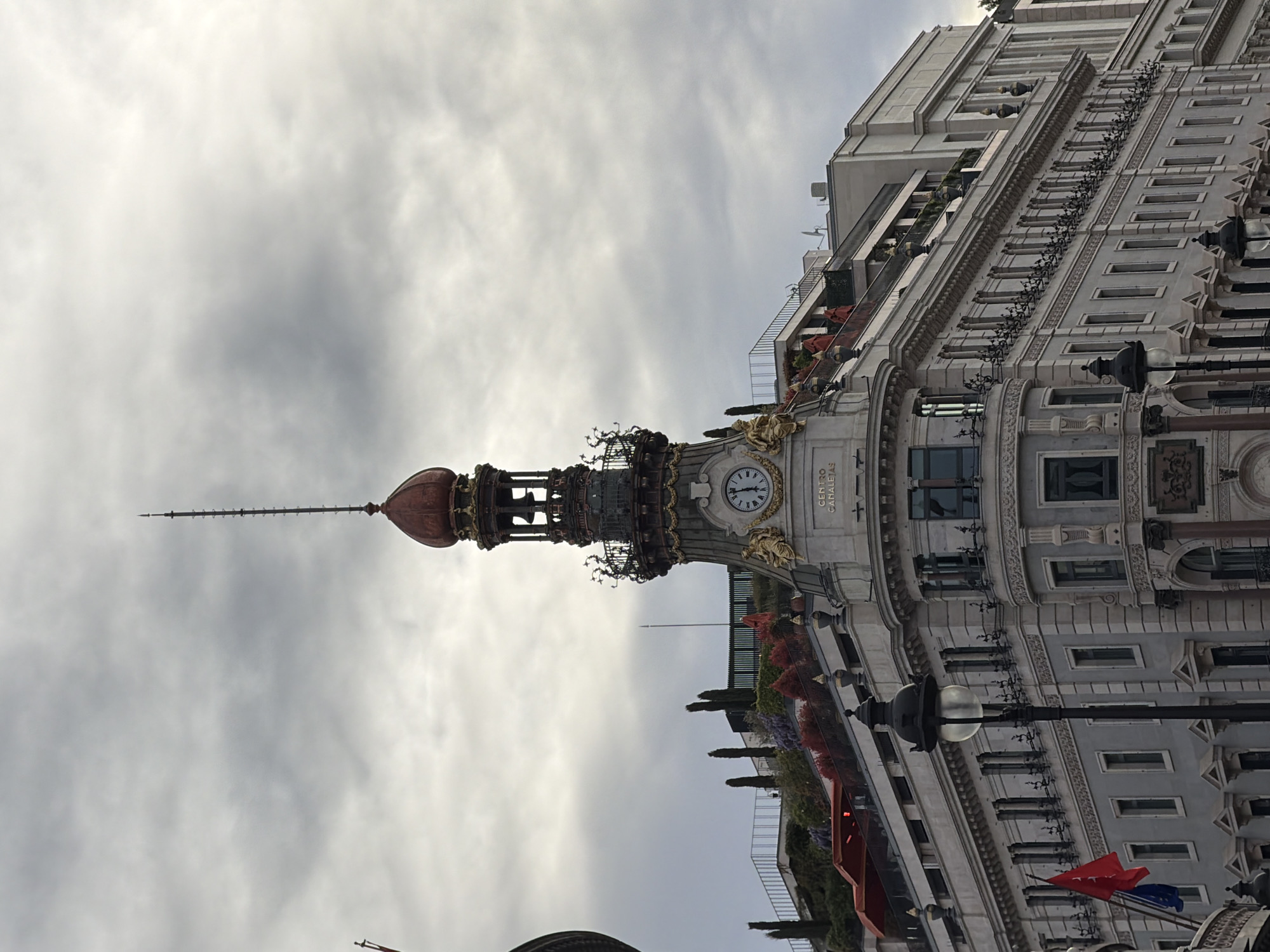 The image shows a portion of the Metropolis Building in Madrid, Spain, viewed from a low angle. The building features a clock tower with a dome and a decorative spire against a cloudy sky. The building's facade is ornate with rows of windows, balconies with plants, and decorative lamps. The inscription "Centro Canalejas" is visible near the clock. Street lamps are present in the foreground.