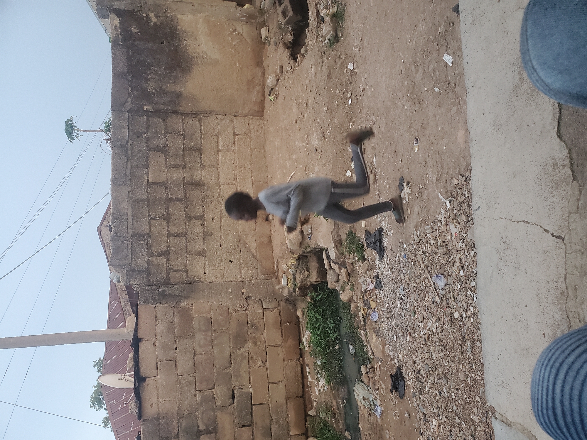A boy is running in an alleyway in Jos, Nigeria. The alley is bordered by brick buildings. There is a drain with green vegetation on the ground. The ground is dirt and small stones, littered with some trash. A building and a utility pole are visible against the sky in the background.