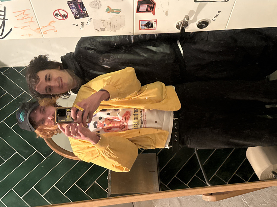 The image depicts a mirror selfie of two individuals, likely male, taken in an indoor public or private space consistent with a restroom or changing room environment. The location is consistent with an urban setting in Tel-Aviv, Israel, indicated by the style of graffiti and cultural elements.
The individual on the left, taking the photograph, has shoulder-length reddish-brown hair and wears a black baseball cap with a green emblem, a septal nose piercing, a yellow bomber jacket, and a white t-shirt featuring a detailed anatomical illustration of a human figure. They are wearing black pants with a multi-eyelet belt. This individual holds a smartphone with a light-colored case and two small stickers (one yellow with text "TIME KILLS") in their right hand, positioned to capture the reflection. A yellow wristband is visible on their left wrist, and rings are on their fingers.
The second individual, positioned to the right of the first, has dark, curly hair and is wearing a black puffer jacket or similar hooded jacket over a light-colored undershirt. This individual is looking towards the camera with a faint smile.
The background on the left features a wall covered in dark green rectangular tiles arranged in a herringbone pattern. A portion of a wooden mirror frame is visible, along with a metal fixture, possibly a hand dryer or dispenser, mounted on the tiled wall. Below the mirror, a white toilet paper holder with a roll is visible. The background on the right consists of a white door or wall panel extensively covered in various graffiti, stickers, and handwritten marks, including what appear to be Hebrew characters. Notable stickers include a red circular 'no entry' symbol and a graphic resembling an old red computer monitor. A chrome door handle and a circular lock mechanism are present on the white panel.