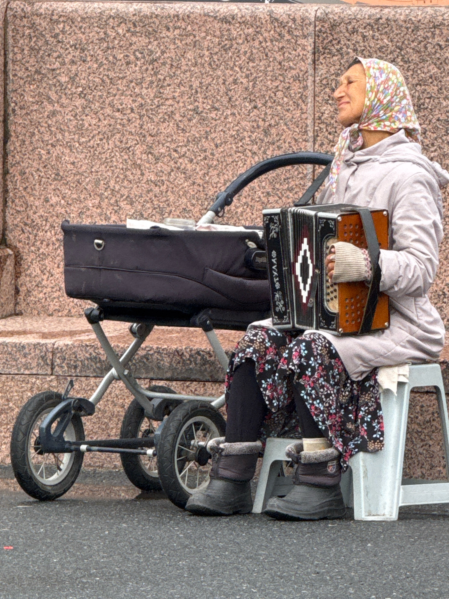 An elderly woman with a headscarf is playing an accordion while sitting on a stool next to a stroller. She is wearing a light-colored jacket, dark patterned leggings, and fur-lined boots. The accordion is brown and black with decorative diamond patterns. The stroller is black with a metal frame and wheels. The background is a textured pinkish-brown wall. The scene appears to be outdoors on a paved surface, possibly a sidewalk or plaza in Veliky Novgorod, Russia, given the architectural style of the background. The lighting suggests it is daytime, possibly overcast.