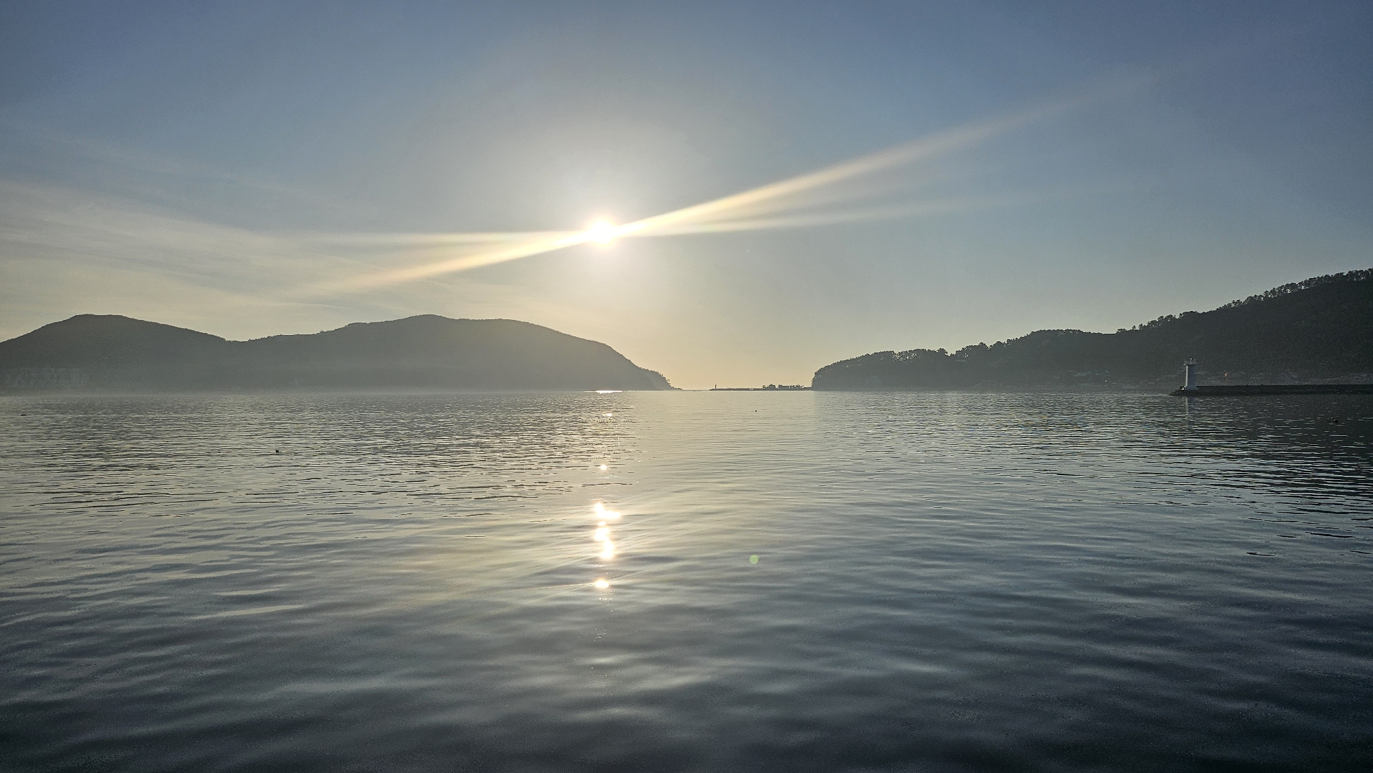 The image depicts a serene seascape in Geoje-si, South Korea, likely during sunrise or sunset. The sun is low on the horizon, casting a warm glow across the water and illuminating the sky with streaks of light. Silhouetted against the bright sky are two hilly landmasses, one on the left and another on the right, covered with vegetation. In the middle distance, a small lighthouse stands on a breakwater, guiding ships into the harbor. The water is calm, with gentle ripples reflecting the sunlight. The overall atmosphere is tranquil and picturesque.