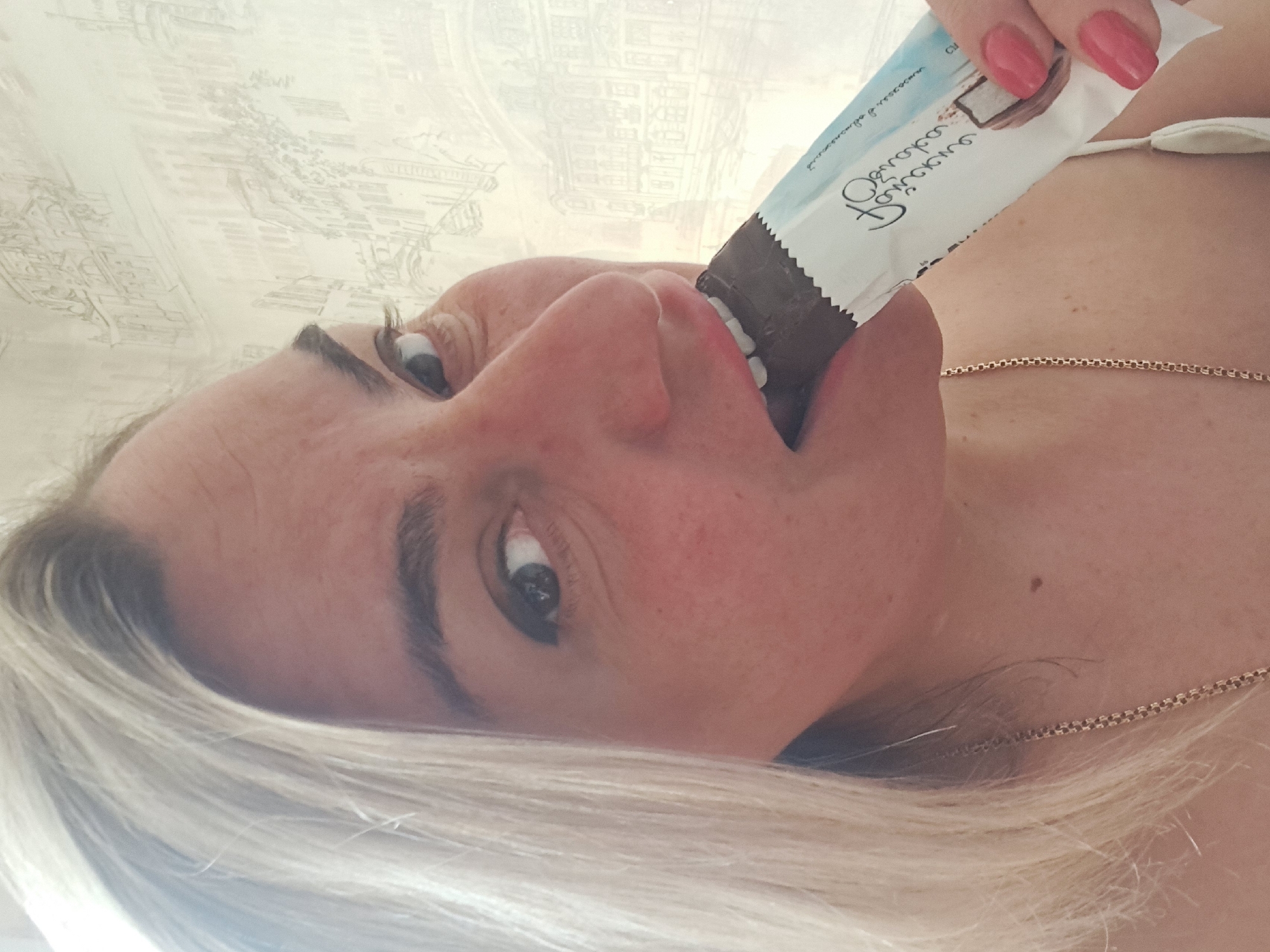 A woman with light blonde hair, wearing a necklace and a top with a white strap, is eating a chocolate bar. Her face is close to the camera. The candy wrapper is white with a picture of a coconut on it, with text in Russian. The background appears to be a light-colored wall with a patterned design. The image appears to have been taken indoors, and there is no indication that it was taken in Elektrostal, Russia.