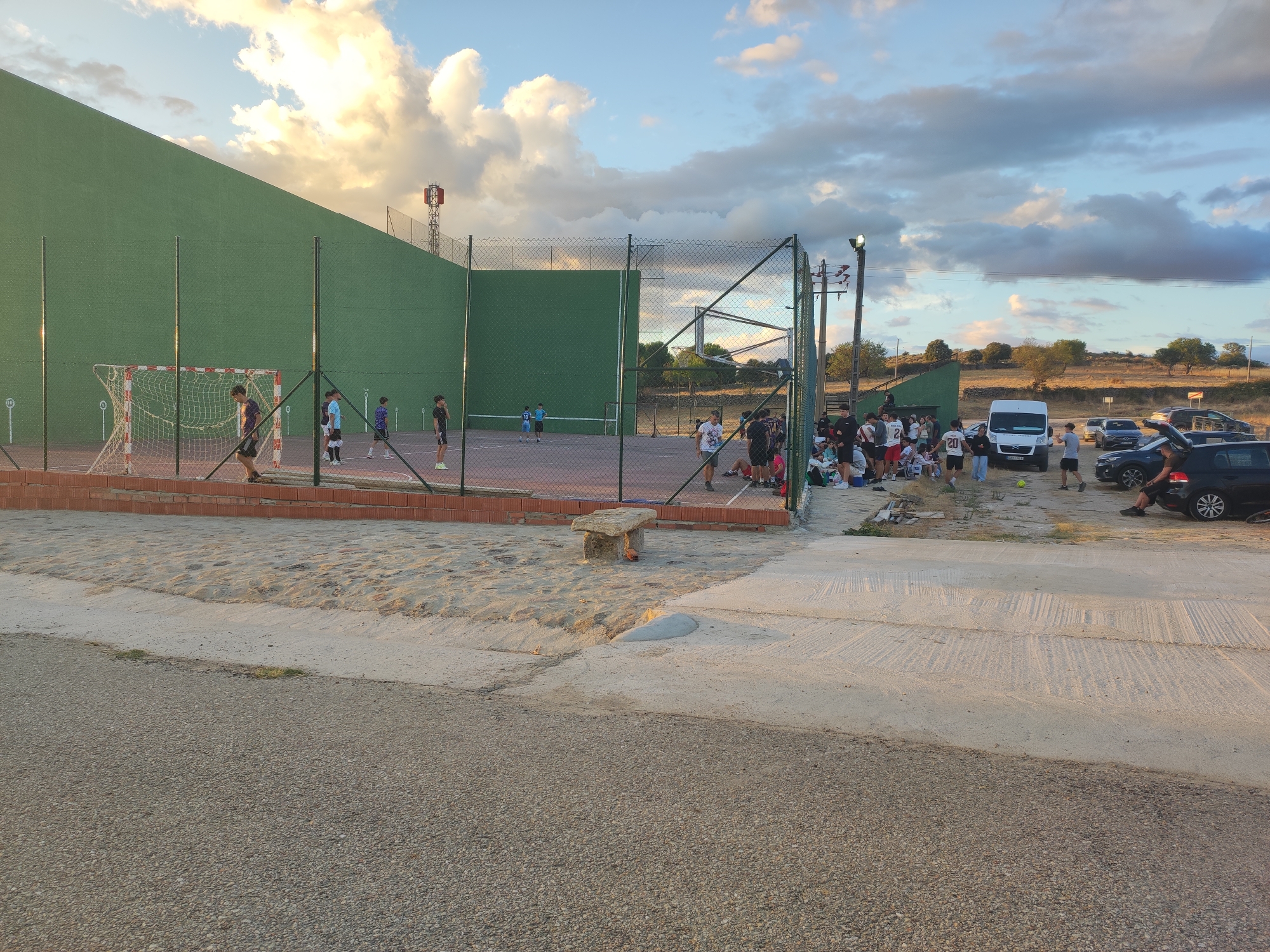 The image depicts an outdoor court facility, likely in Sobradillo, Spain, during daylight hours. The court appears to be multi-purpose, with a large green wall, a net, and goalposts suggesting it can be used for sports like handball or perhaps a simplified version of soccer or tennis. Several individuals, predominantly young men and some women, are scattered around the premises. Some are actively engaged in sports on the court, with at least two appearing to be playing. Others are gathered in groups outside the court, socializing and watching. A white van and several cars are parked nearby, indicating vehicular access and a likely gathering of people. The environment is somewhat arid, with sparse vegetation visible in the background and a sandy area adjacent to the court. The sky is partly cloudy, with dramatic cloud formations illuminated by the sun, suggesting it might be late afternoon or early evening.