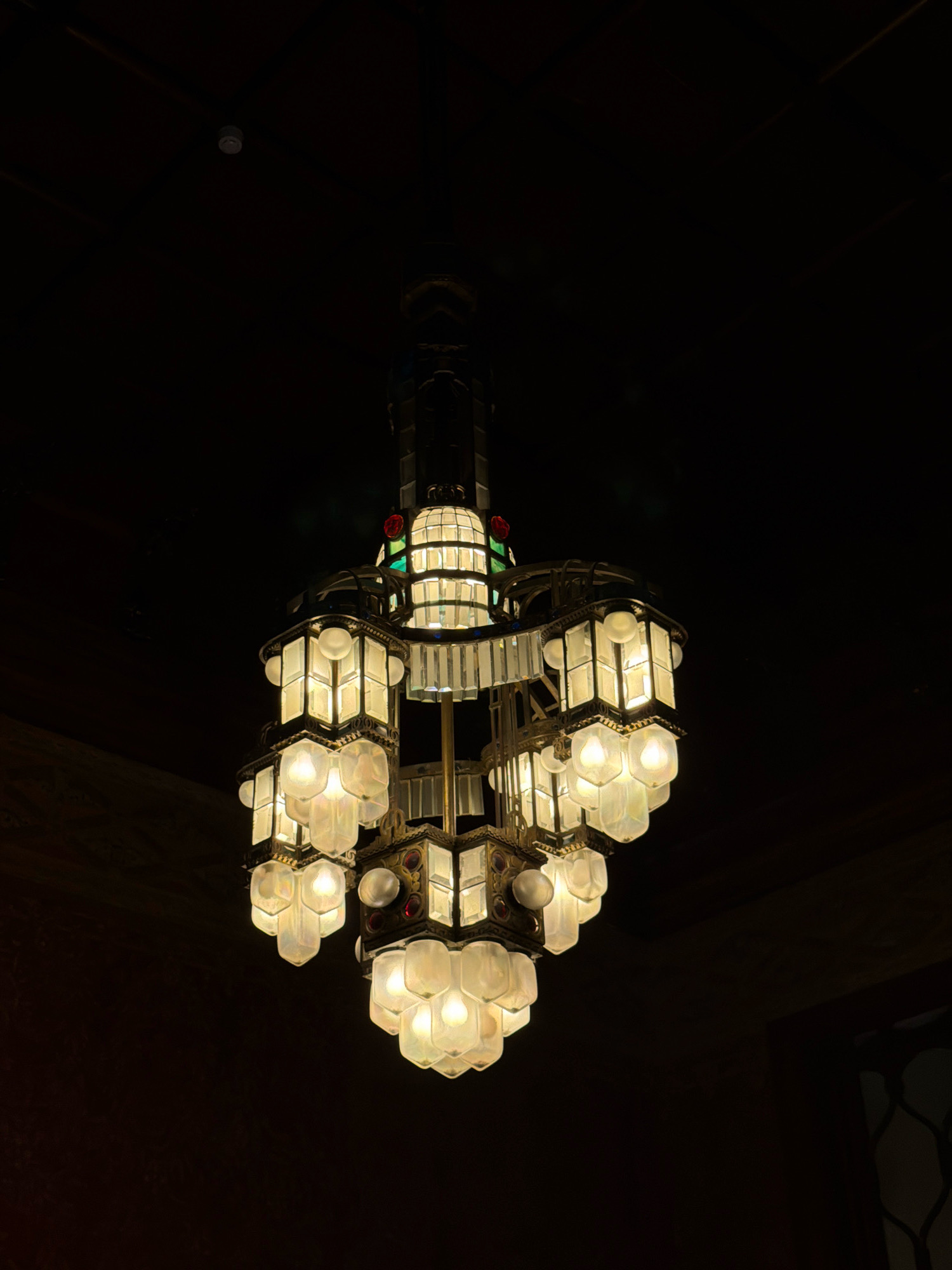 An ornate, multi-tiered chandelier is the central subject of the image, suspended against a dark background. The chandelier is made of brass and features several hexagonal light fixtures with frosted glass panels, illuminated from within, emitting a warm, yellow glow. Some of the fixtures also incorporate faceted clear glass and small, colored glass accents, appearing to be red and green. The chandelier's structure is complex, with vertical elements and cascading light clusters. The background is predominantly dark, suggesting an indoor setting with low ambient light. The upper portion of the image shows a dark ceiling with some faint horizontal lines, possibly beams or architectural features. The lower portion of the image transitions into a dark, textured wall, with a hint of decorative grillwork on the far right. The image appears to be taken from below, looking up at the chandelier. There are no people visible in the image. The overall impression is of a historical or decorative lighting fixture in an dimly lit interior space. The location is identified as the Grand Hotel, San Pellegrino Terme, Italy, known for its Art Nouveau architecture.