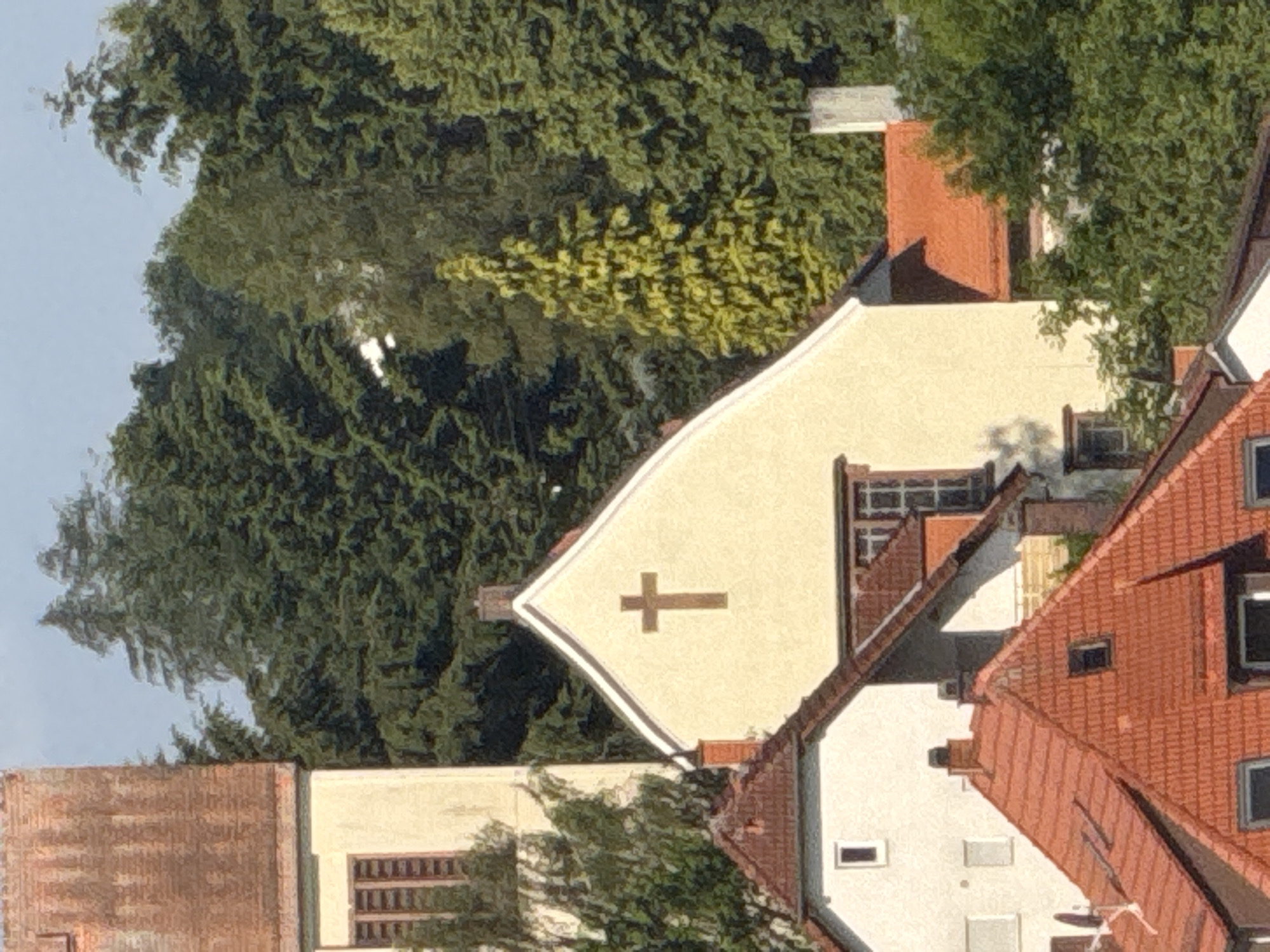 The image shows a residential area in Strümpfelbrunn, Germany, with several buildings and dense trees in the background. A prominent feature is a light-colored building with a white arched facade, prominently displaying a brown cross. Adjacent to it, to the left, is a taller building with a red-tiled roof and a cream-colored facade, featuring a tall, narrow window. In the foreground, several red-tiled roofs of houses are visible, some with visible chimneys and windows. The environment appears to be a sunny day, with shadows indicating the presence of sunlight. The background is dominated by a thick canopy of various green trees, suggesting a natural or semi-natural setting. No people are visible in the image.