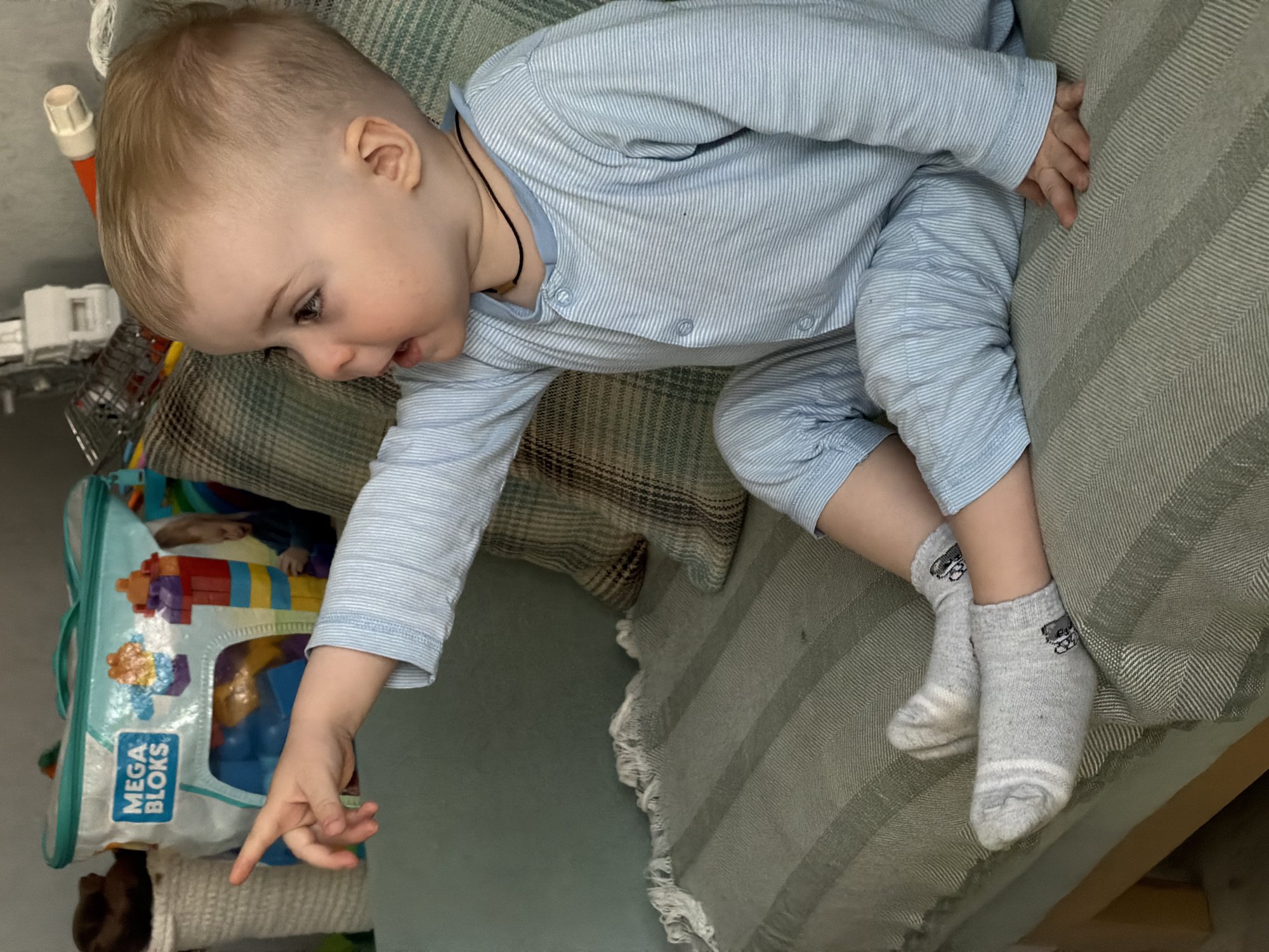A young child, likely a toddler, lies on a striped, green and beige sofa. The child wears a light blue, striped one-piece garment and grey socks with a small cartoon car on each. The child is fair-skinned with blonde hair and is turned towards the left of the frame, pointing down with their right index finger. Beside the child on the sofa is a shopping cart, a white object with an orange handle, and a clear bag with the words "MEGA BLOKS" visible.
There is no indication of Votkinsk, Russia in this image.