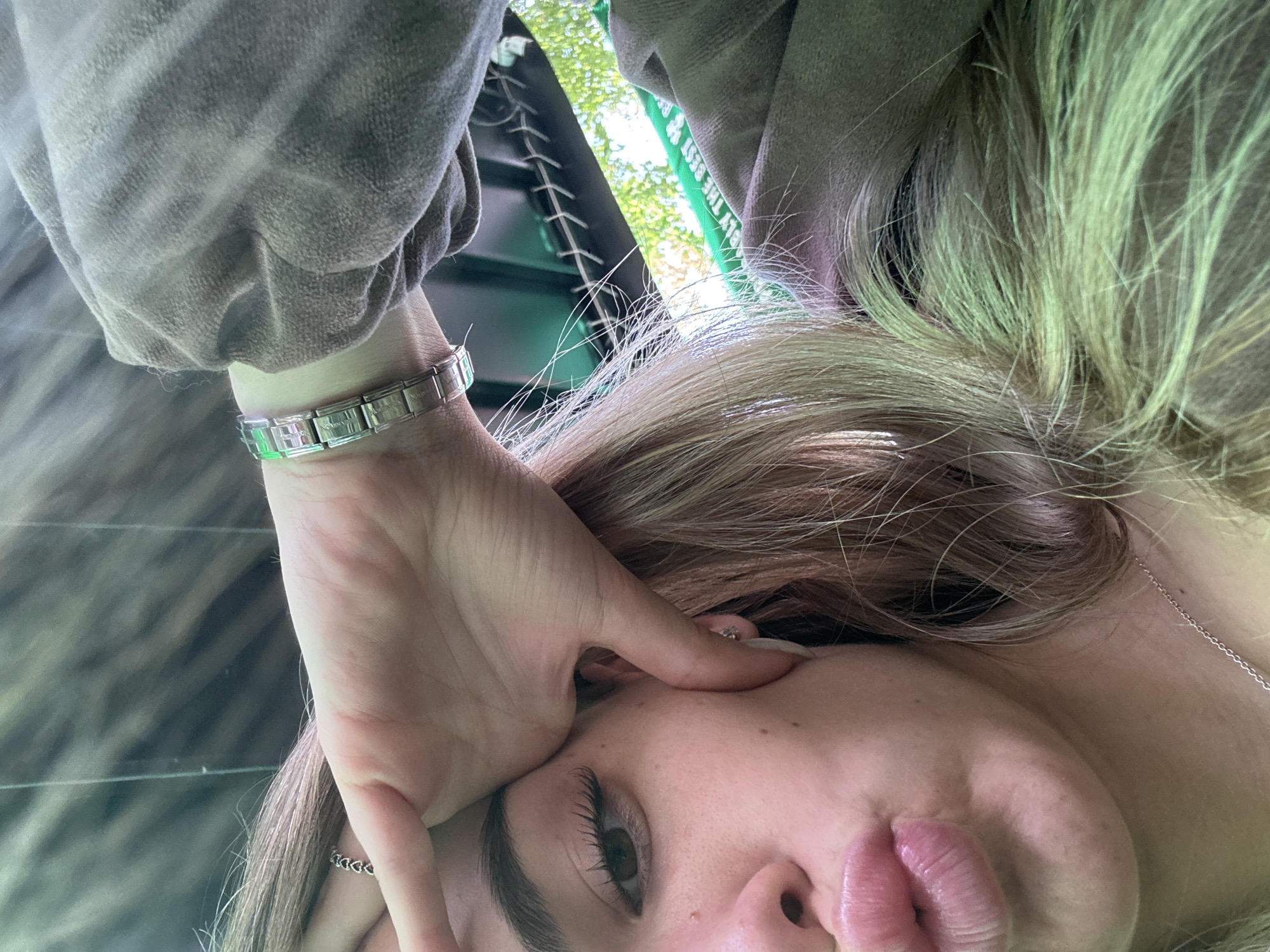 A close-up portrait of an individual, likely female, is captured from an unusual, possibly overhead or angled, perspective. The subject has long, light brown and blonde hair and is wearing a textured, grey long-sleeved garment, possibly a sweatshirt.
Her left hand, adorned with a silver modular link bracelet, rests against her face near her temple. A thin silver chain necklace is visible around her neck. Her eye is open, showing a dark iris and long eyelashes, and her lips are slightly pursed.
The background is out of focus, revealing hints of an outdoor setting with green foliage and a dark structure, possibly metal or plastic. A green strap with partially visible white text can be seen in the background, appearing to read "PLY THE BEST W". The bright, natural lighting indicates the photograph was taken during the daytime. The overall impression is a casual, intimate shot, taken in Sofia, Bulgaria.