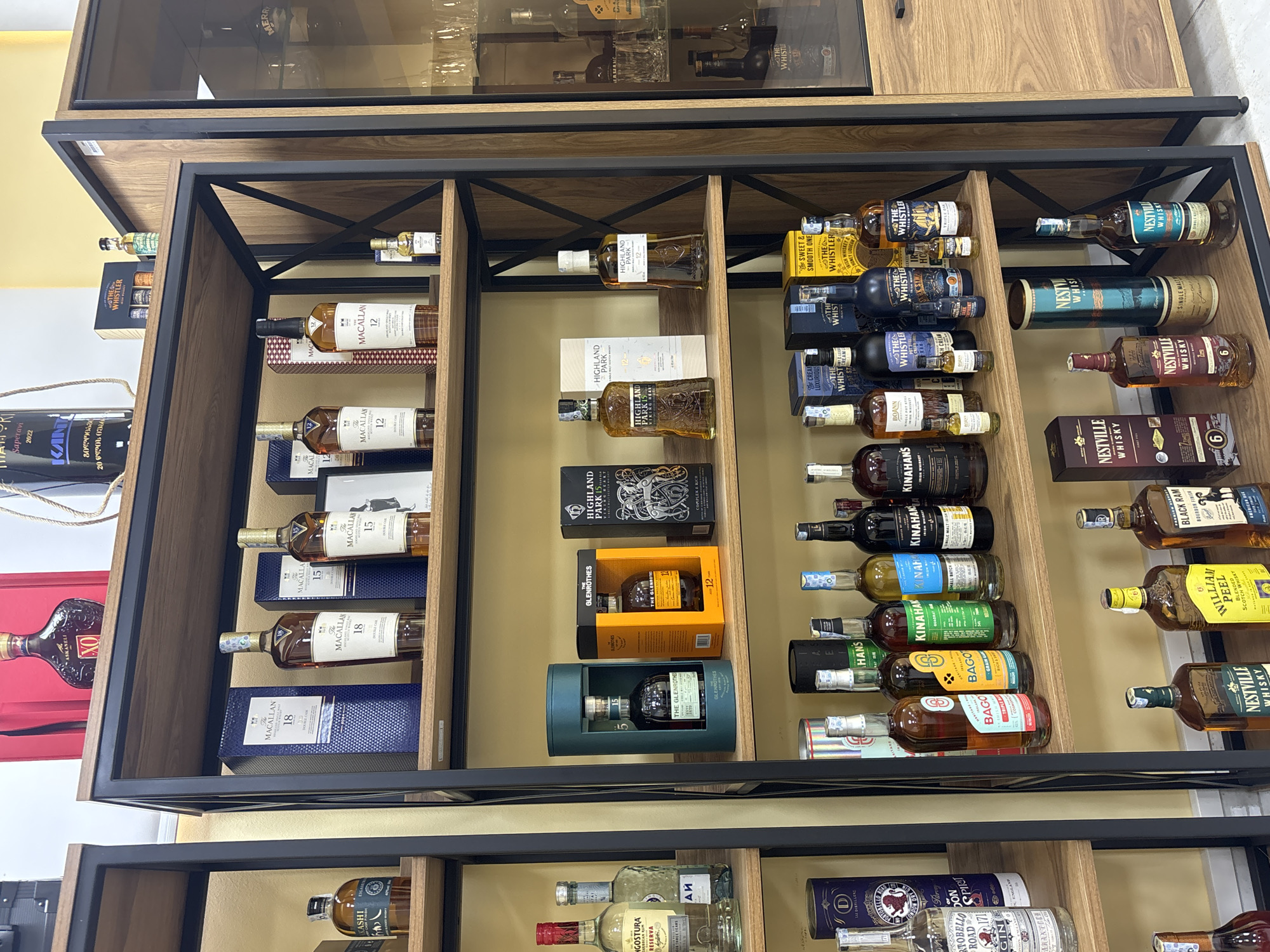 The image shows a retail display of various alcoholic beverages, primarily whiskeys, arranged in wooden shelving units with a black metal frame. The brands include The Macallan, The Glenrothes, Highland Park, The Whistler, Kinahan's, Boann, Nestville Whiskey, William Peel, Black Ram and Bago. Some of the bottles are in boxes or tubes. The shelves are well-stocked with a variety of whiskeys, suggesting a store specializing in alcohol sales. The text "Saperavi 2022" and other Georgian words on a bottle located towards the left side of the image, combined with the fact the image is showcasing various whiskey brands, suggests the photo was taken in Tbilisi, Georgia.