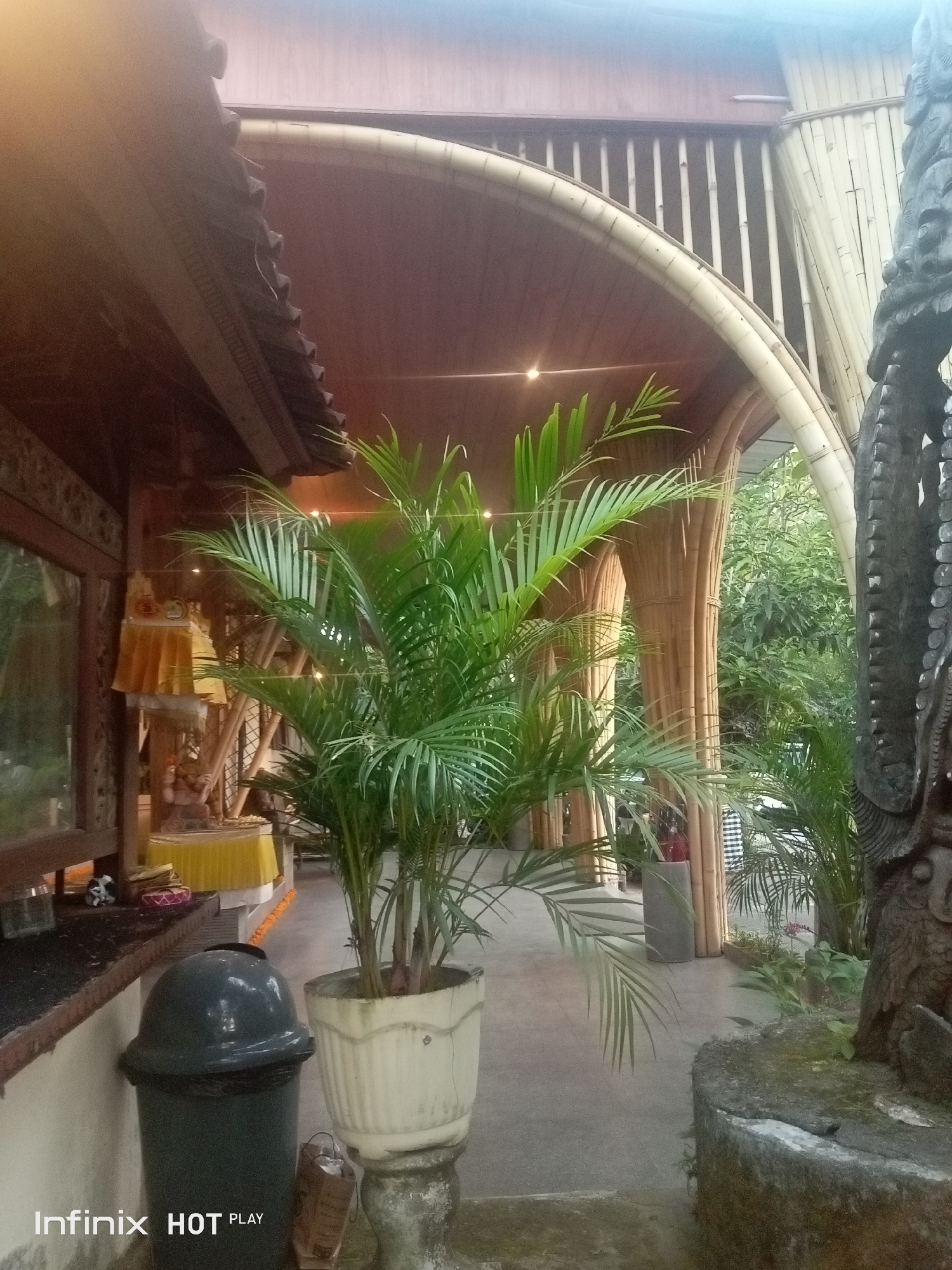 The image depicts an outdoor covered walkway with a rustic, natural aesthetic. The structure features a distinctive arched bamboo roof supported by vertical bamboo poles. To the left, a wooden building with decorative carvings is visible, alongside a large window. Inside this building, yellow fabric draped over what appears to be a shrine or altar can be seen, along with a small statue. In the foreground, a potted palm plant with lush green fronds stands prominently, situated next to a dark grey trash can and a white planter on a pedestal. A carved stone statue, partially visible on the right side of the frame, adds to the cultural ambiance. The ground is paved with concrete. The overall impression is that of a serene, possibly spiritual or traditional, outdoor space in Ubud, Indonesia, characterized by natural materials and decorative elements.