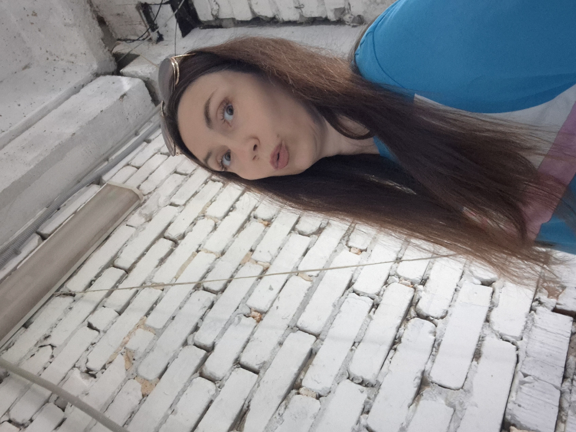 A medium shot captures a Caucasian woman with long brown hair and sunglasses perched on her head, posing for a selfie against a white brick wall. Her eyes are wide, and her lips are pursed in a playful expression. She is wearing a blue t-shirt with a white and pink stripe across the chest. The background shows a textured white brick wall with visible mortar. Above and to the right of the woman, a portion of a concrete ceiling with exposed wiring and conduit is visible. The scene appears to be indoors, possibly in an unfinished or industrial space, located in the city of Chaikovsky, Russia.