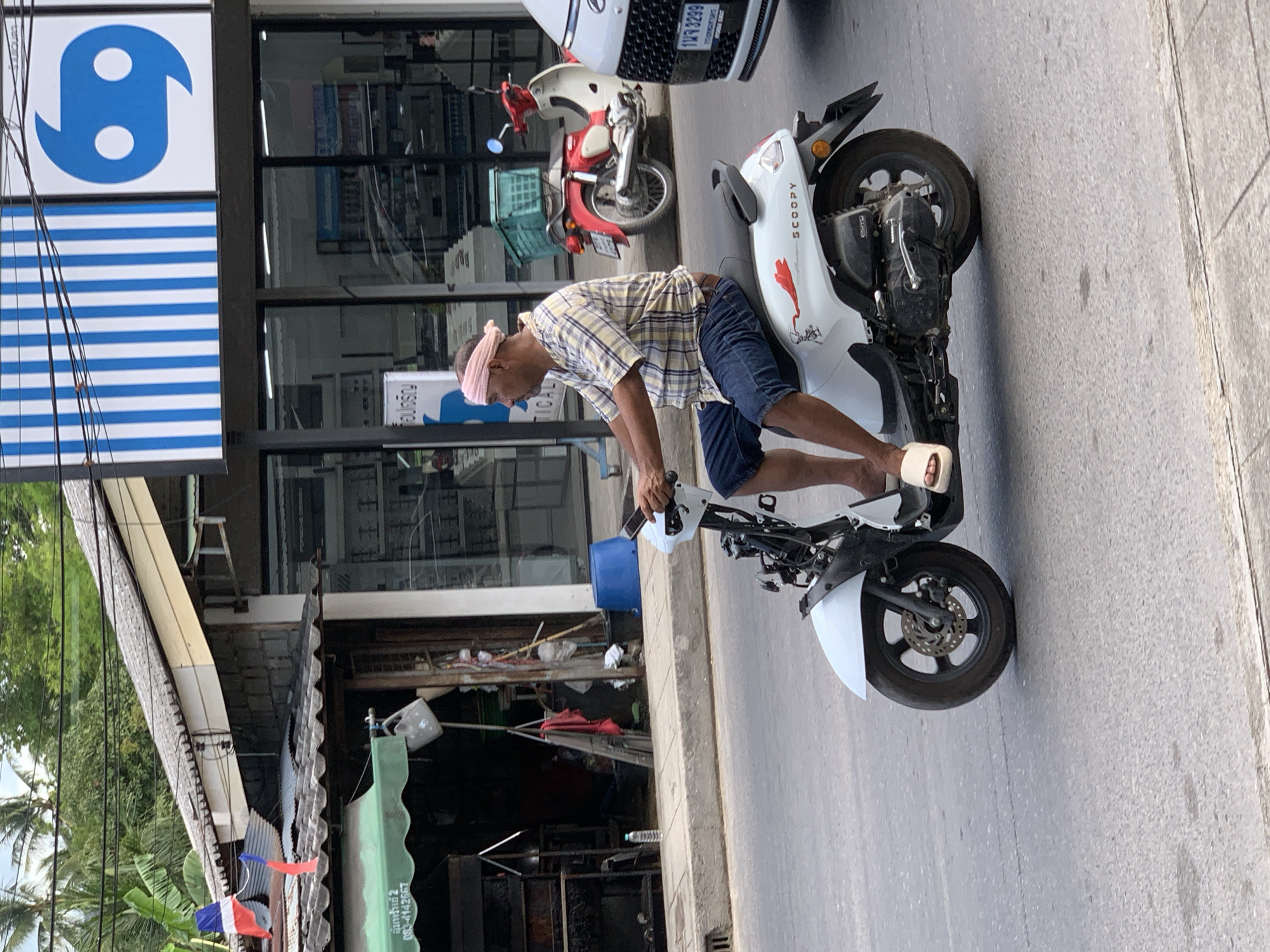 A man is riding a white Honda Scoopy scooter on a paved street in the city of Ko Samui District, Thailand. He is wearing a plaid shirt, blue denim shorts, and light-colored sandals. A pink cloth is tied around his head. The scooter has a red flower decal on the rear fender. In the background, there is a building with a blue and white striped awning and a blue logo. A red Honda motorcycle is parked in front of the building. Other elements visible include a sidewalk, trees, power lines, and a white car on the right side of the frame.