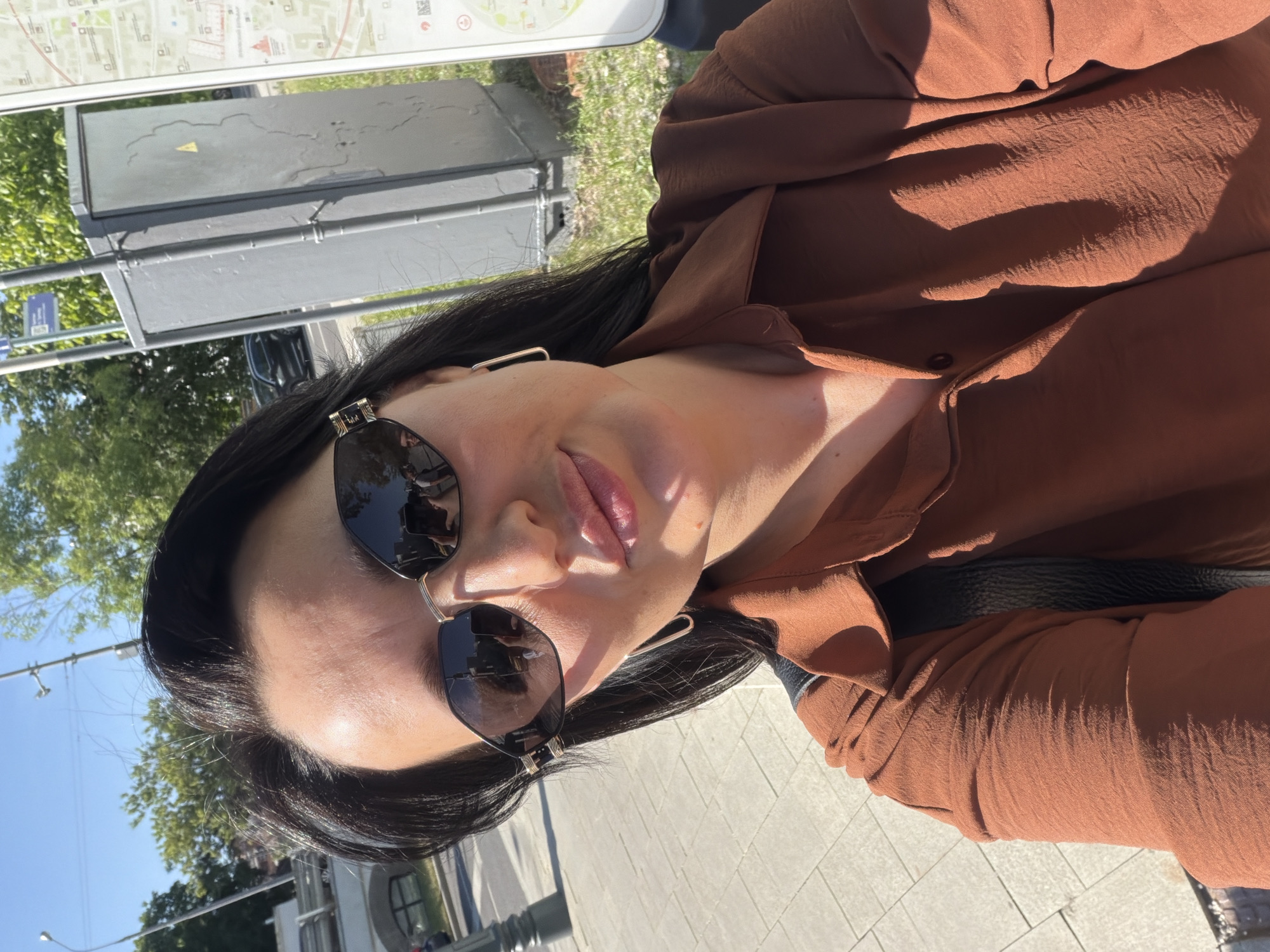 The image displays a waist-up, slightly angled selfie of a woman in an outdoor urban setting under bright sunlight. The woman has dark hair styled away from her face and wears oversized octagonal sunglasses with dark lenses and gold-colored accents on the temples. Her lips are lightly colored and slightly pursed. She wears rectangular gold-colored hoop earrings. Her attire consists of a rust-colored or burnt orange long-sleeved button-down shirt made of a textured fabric. A black strap, likely from a shoulder bag, is visible over her left shoulder. Reflections in her sunglasses show a hand holding a smartphone, confirming the selfie nature of the shot, along with distorted outlines of an urban environment and figures.
To the woman's left, light-colored rectangular pavers form a sidewalk. In the background on the left, a segment of an arched bridge structure in light-colored stone or concrete is partially visible, with a black lamppost, distant green trees, and dark-colored vehicles on a street. To her right, a tall, grey metallic utility box with a yellow triangular hazard symbol on its side stands alongside a white public information stand. This stand displays a detailed city map with Cyrillic text, prominently featuring "МОСКВА" (Moscow) at the top, indicating public transport routes, a red Kremlin-like icon, and subway station markers. A blue rectangular street sign on a pole in the upper right background explicitly identifies the location as "КРАСНОПРЕСНЕНСКАЯ НАБЕРЕЖНАЯ" (Krasnopresnenskaya Embankment). The overall scene depicts a sunny day on Krasnopresnenskaya Embankment in the city of Moscow, Russia.