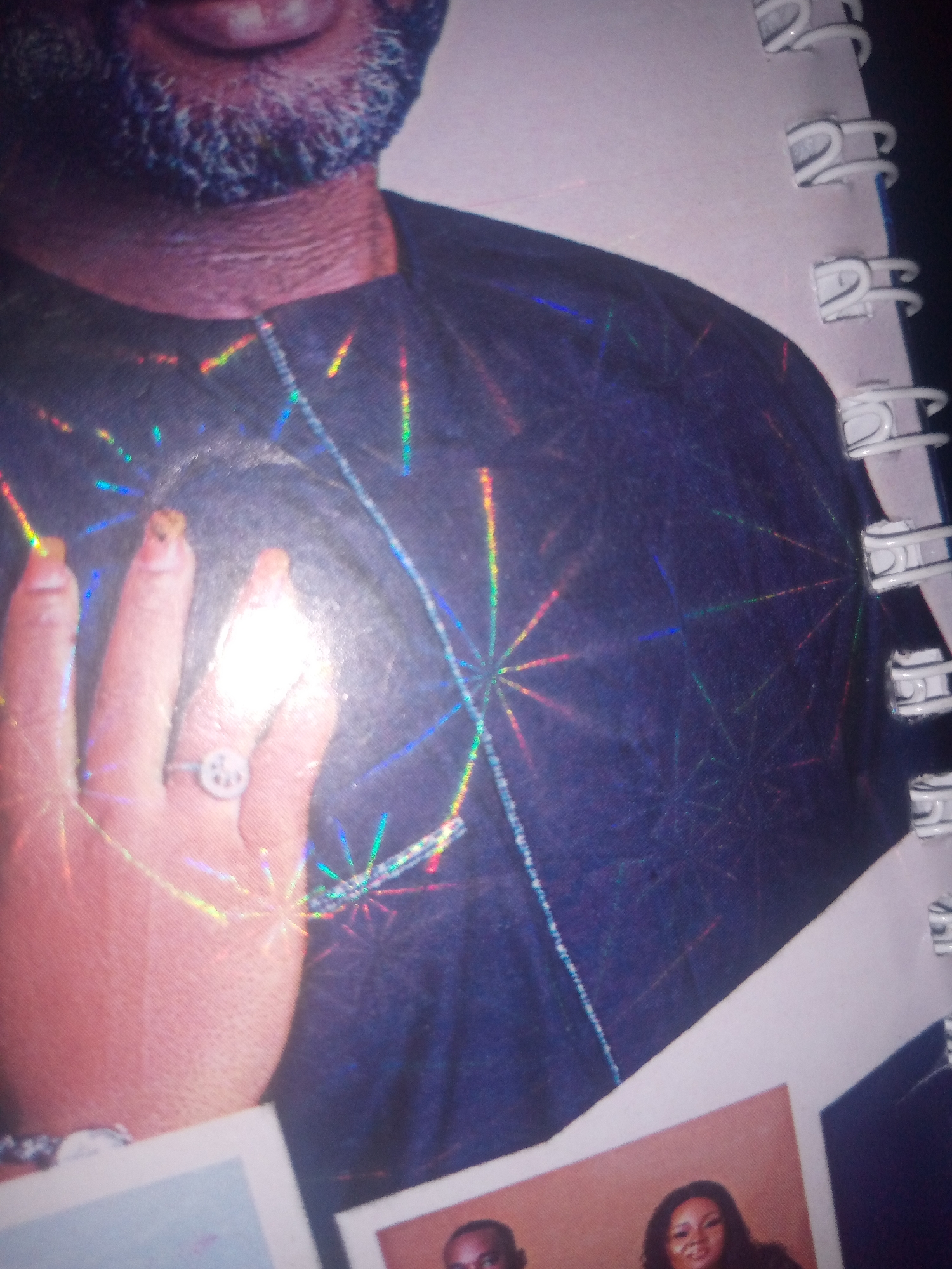 The image is a close-up, angled photograph of a person's left hand and arm, with a portion of their torso visible. The hand is wearing a ring with a circular design. The individual is wearing a dark, possibly navy blue, shirt that has a holographic, starburst-like pattern on it, with iridescent streaks of color reflecting the light. The background appears to be a notebook or binder with white spiral binding on the right side. There are also parts of two photographs visible at the bottom of the image, showing the heads of two individuals. The lighting is strong and creates bright highlights, particularly on the hand and shirt. The scene is not specific to any identifiable location in Jalingo, Nigeria, as there are no contextual clues such as landmarks or cultural elements.
