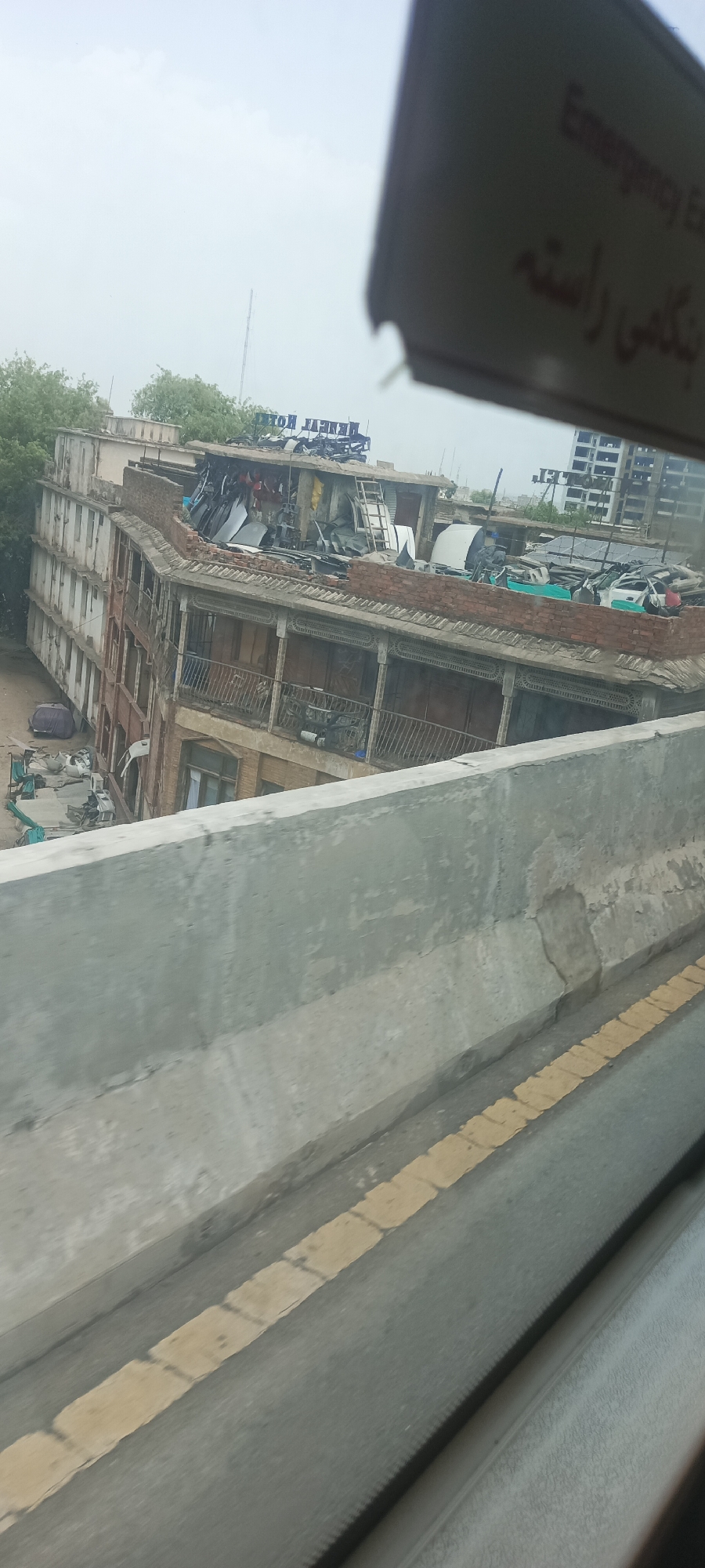 The image depicts an urban scene in Alam Khan Kalay, Pakistan. The central subject is an old, multi-story building with a distinctive curved facade. The building appears to be in a state of disrepair, with damage to its roof and upper stories. The roof is cluttered with various objects, including what appear to be car parts, scrap metal, tarps, and a ladder, suggesting it may be used for storage or dismantling. The balconies on the lower floors feature intricate wooden latticework.
To the left of the building, there is some greenery, indicating the presence of trees. In the foreground, a concrete barrier runs diagonally across the frame, with yellow road markings visible on the asphalt road beyond it. The sky is overcast, suggesting cloudy weather. The overall impression is one of a bustling, possibly working-class, urban environment.
A sign is partially visible at the top right of the image, tilted and partially obscuring the sky. The visible text on the sign appears to be in English and potentially another language, possibly Urdu or Arabic. The English text includes "Emergency Ex" and what looks like "ring". Below this, there is text in a script that appears to be Arabic or Urdu, with the word "راسته" being discernible, which translates to "route" or "way" in Urdu. A sign on the building itself has text that appears to read "HOTEL BENEKI" or similar, though it is reversed and partially obscured.
No people are clearly visible in the image, but the presence of a parked vehicle to the left of the building suggests some activity. The time of day is difficult to determine precisely due to the overcast sky, but it appears to be daytime. The scene conveys a sense of everyday life and industry within a Pakistani urban setting.