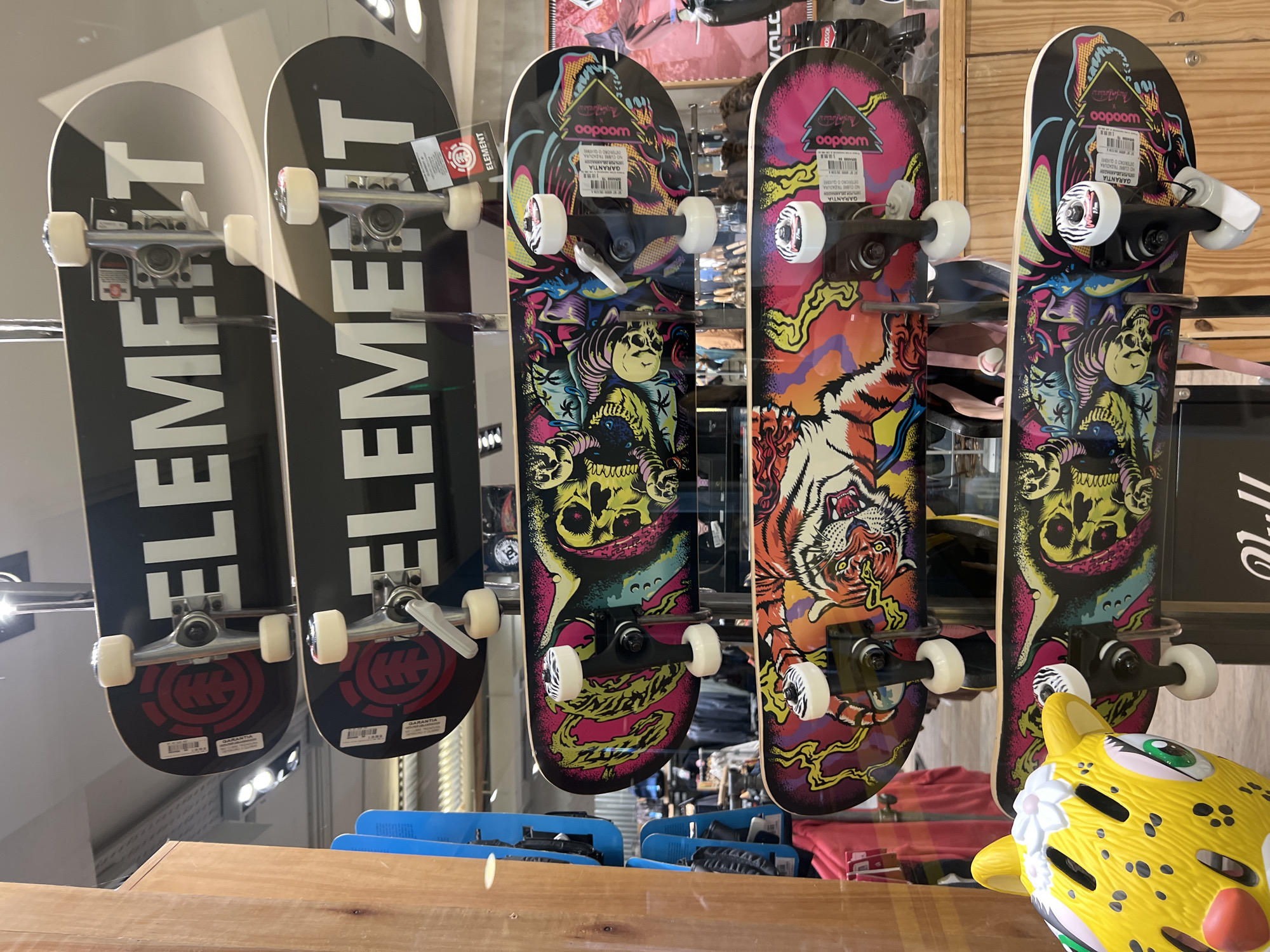 The image shows a display of five skateboards in what appears to be a retail store. The skateboards are arranged in a row, with two black skateboards on the left, each bearing the word "ELEMENT" in large white letters and the Element logo on the bottom, and three more colorful skateboards to the right of them. These colorful boards feature more complex graphics, with one showing an orange and yellow tiger, and the other two showing a skull with a snake. All the skateboards have trucks and wheels attached. Tags are visible on the skateboards, suggesting they are new and for sale.
In the foreground, a yellow helmet designed to look like a leopard's head is partially visible.
The skateboards are behind a clear display window, through which reflections of the store's interior and possibly the surrounding environment are visible. The objects in the background appear to be merchandise within the shop, including clothing and other retail items.
The text present in the picture includes "ELEMENT", "woodoo", "GARANTIA", and "VOLC". A sign saying "KOSSOK" is visible. The location is suggested as the city of Buenos Aires, Argentina.