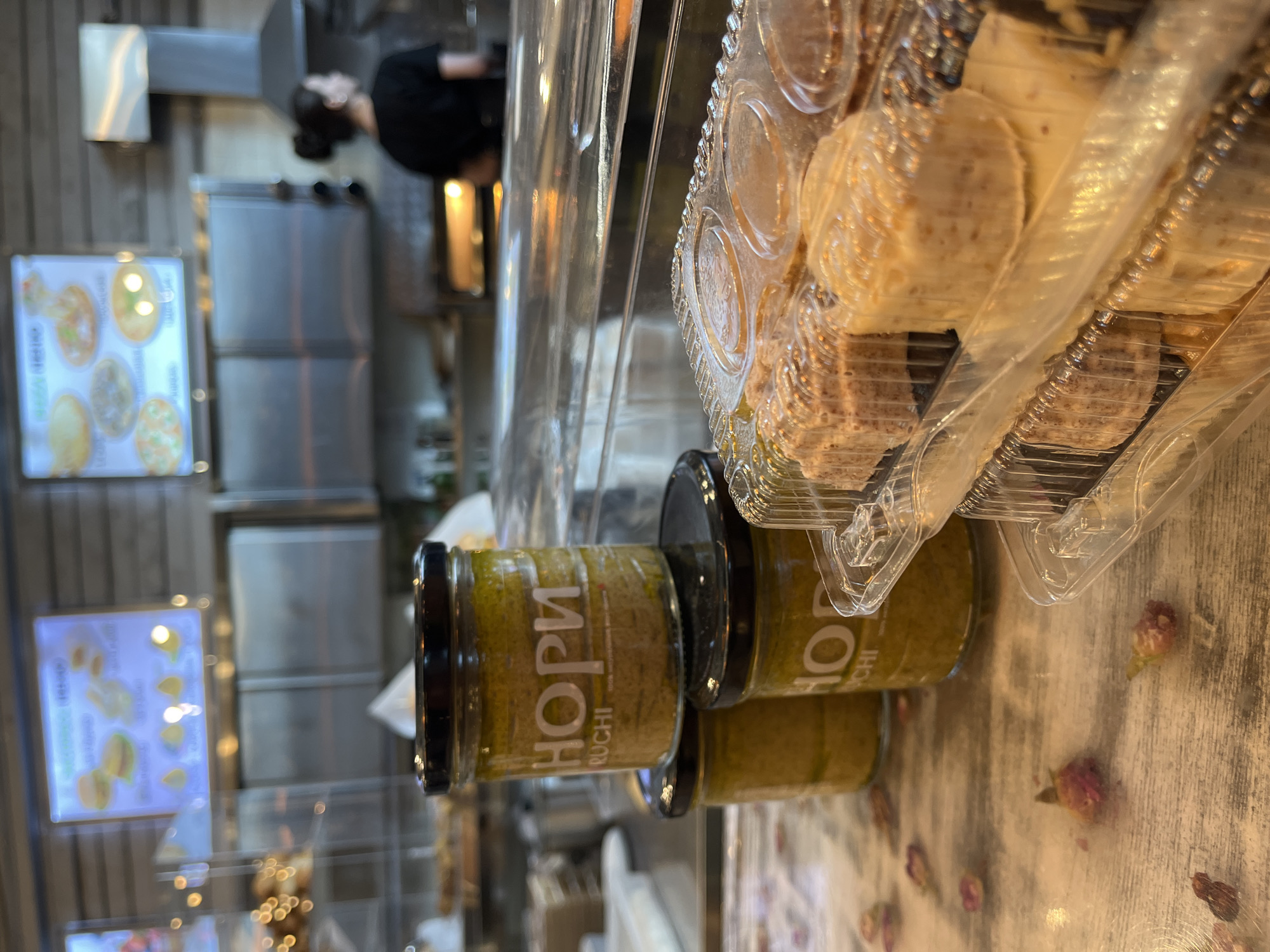 The image shows a close-up of a display counter in what appears to be a food establishment. In the foreground, three glass jars are stacked, with the top jar clearly displaying the "HORI RUCHI" label. To the right, a transparent plastic container holds several portions of what looks like dessert or pastry items. In the background, a person wearing a black shirt is visible, looking towards the right, and kitchen equipment, including ovens and a ventilation hood, suggests an operational food preparation area. A menu board displaying food images is mounted on the wall. The overall setting seems to be a casual dining or take-out restaurant. The presence of dried rose petals scattered on the table adds a decorative element to the scene.