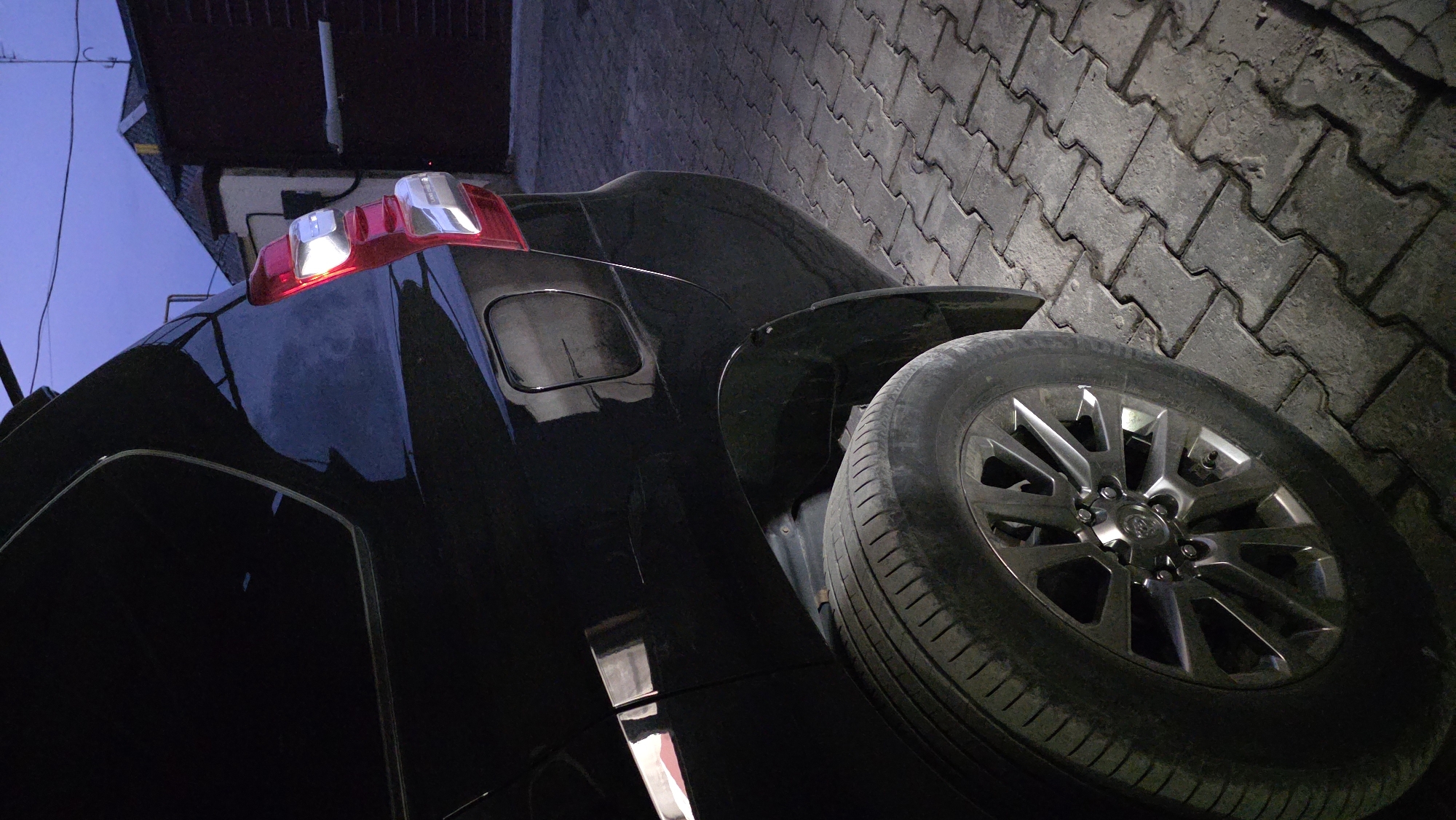 The image shows the rear quarter panel and wheel of a black SUV parked on a paver stone surface. The vehicle's taillight is illuminated. In the background, a brick building with a dark roof and some overhead wires are visible. The sky appears to be twilight. The scene is set in the city of Chuy, Kyrgyzstan.