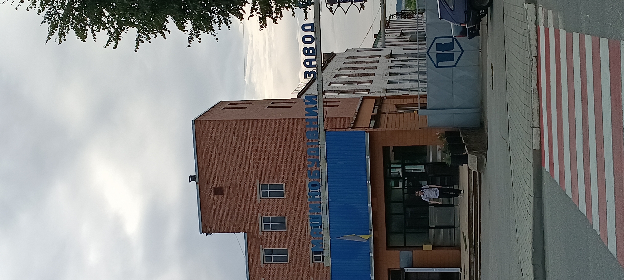 The image depicts the entrance of a factory in the city of Kamianka, Ukraine. A man wearing a collared shirt and dark pants is walking towards the viewer from inside the building. Above the entrance, large blue letters spell out "МАШИНОБУДІВНИЙ ЗАВОД" which translates to "MACHINE-BUILDING FACTORY". The factory building is constructed of red brick and has several windows. To the right of the building, a section of a white building with numerous windows is visible. A grey metal fence with a blue logo is in the foreground, partially obscuring a dark colored car. A pedestrian crossing with red and white stripes is in the lower part of the image. The sky is overcast with clouds.