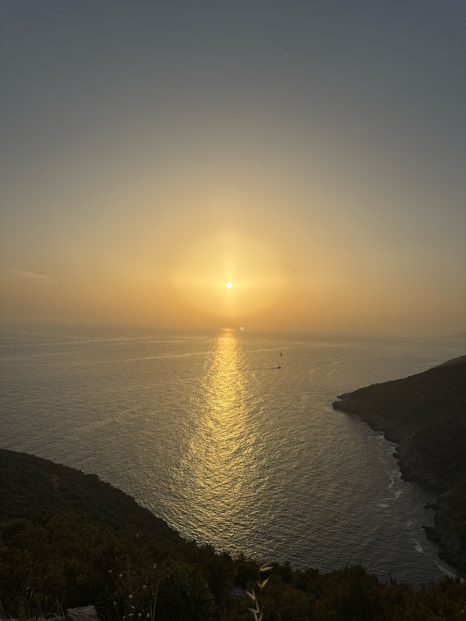 The image captures a serene sunset over the Adriatic Sea, with the sun positioned centrally in the frame, casting a golden reflection across the water. The sky transitions from a pale blue at the top to a warm yellow glow near the horizon, softened by a slight haze. Two sailboats are visible in the distance on the sea. The foreground features lush green hillsides, densely covered with trees and foliage, descending towards the water. One of these hillsides forms a curved coastline on the right side of the image, where rocky outcrops meet the sea. The overall impression is one of natural beauty and tranquility. The location is identified as Zagora, Montenegro.