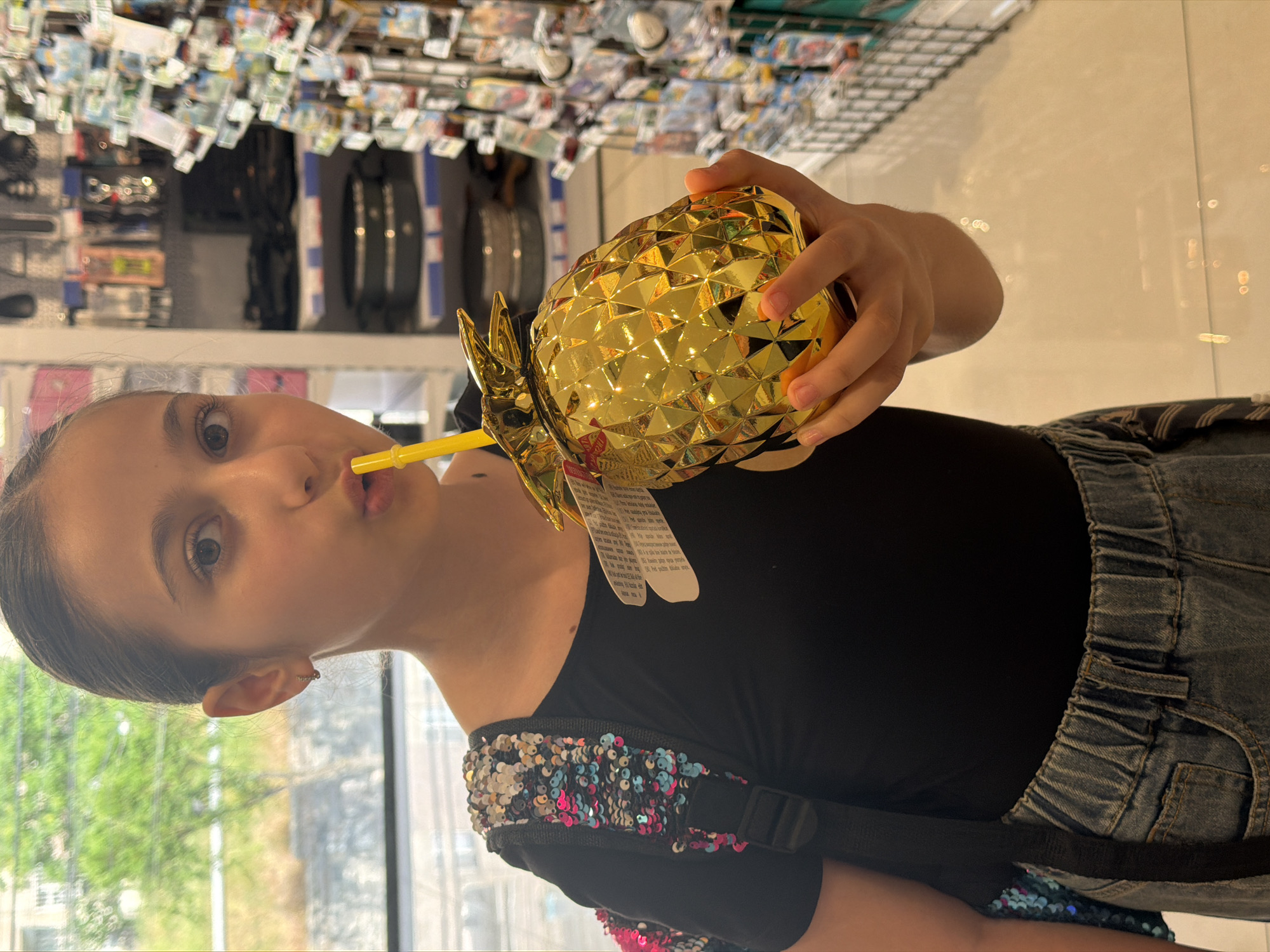 A young girl with dark hair pulled back is holding a gold pineapple-shaped cup with a yellow straw, taking a sip from it. She is wearing a black top and denim shorts, with a backpack adorned with colorful sequins on her left shoulder. The background suggests an indoor retail environment, possibly a shop or store, with shelves stocked with various items. The lighting is bright, and the overall scene is casual. The image was captured in Yerevan, Armenia.