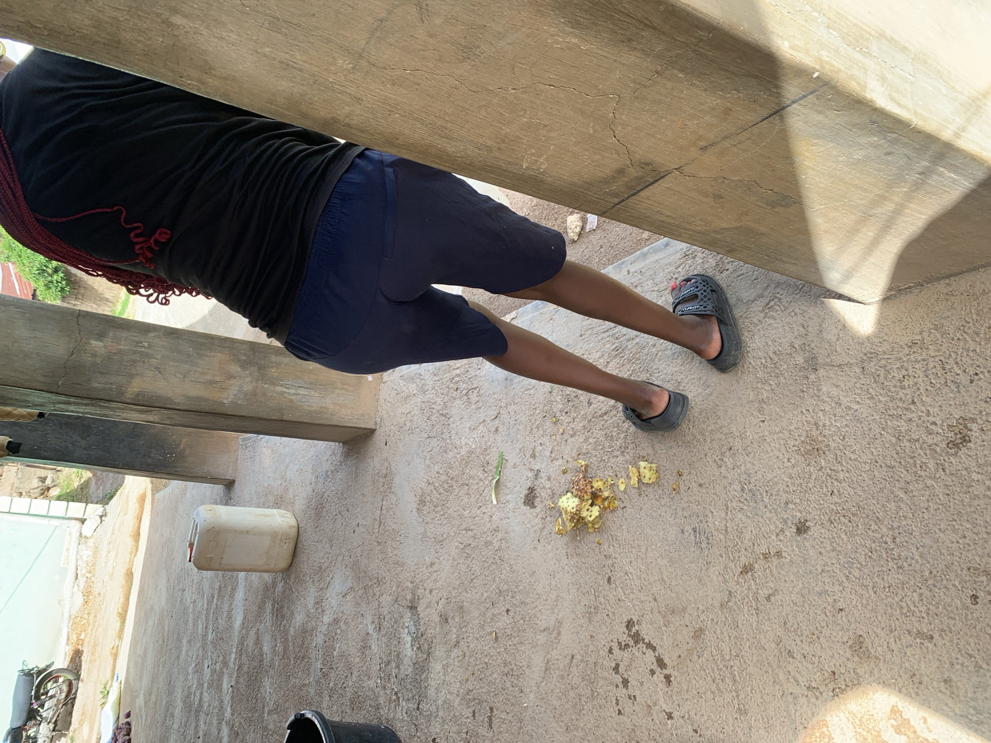 A person wearing a black t-shirt and navy blue shorts stands near a concrete pillar. Their back is to the camera, and their dark, braided hair is visible. The person is wearing dark gray slide sandals. On the concrete ground to the left, there is a pile of what appears to be discarded food scraps, possibly from a pineapple, along with a white plastic container and a black bucket. In the background, a motorcycle is parked, and a light blue building is visible, consistent with a scene in Jos, Nigeria. The overall environment suggests an outdoor, possibly residential or commercial, area.