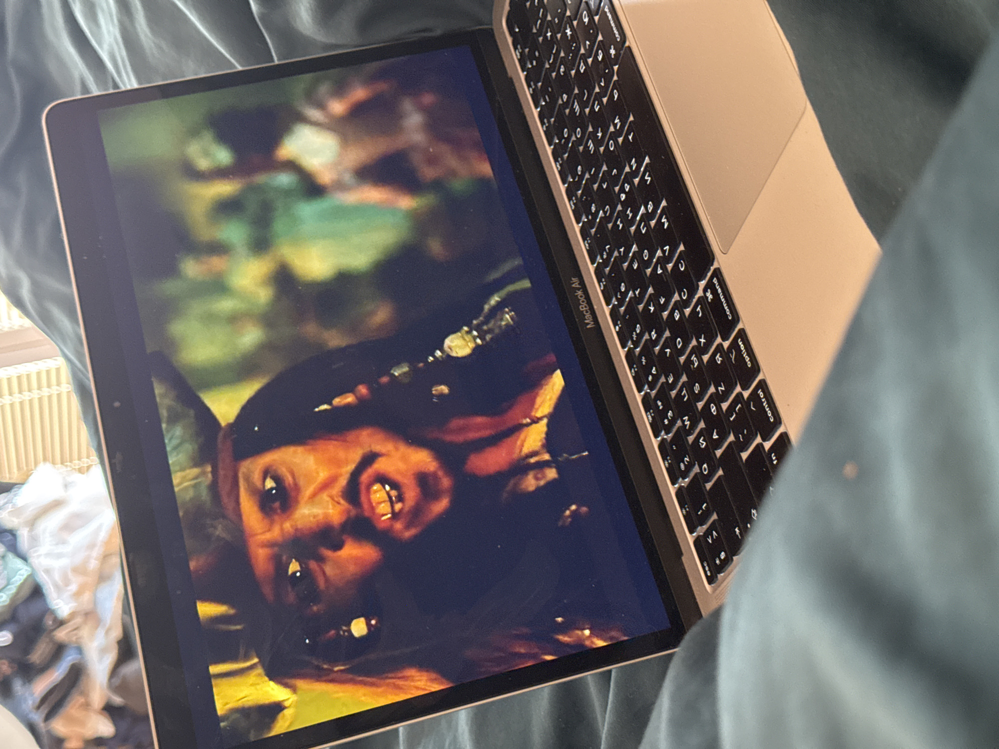 A partially opened gold-colored MacBook Air laptop is positioned on top of teal-colored bedding. The laptop screen displays a slightly upside-down image of what appears to be a character from the movie "Pirates of the Caribbean". The laptop's keyboard, which is partially visible, features Cyrillic characters alongside the standard English alphabet. The setting is indoors, suggested by the bedding and laptop. There is no evidence to support that it is in Budva, Montenegro.