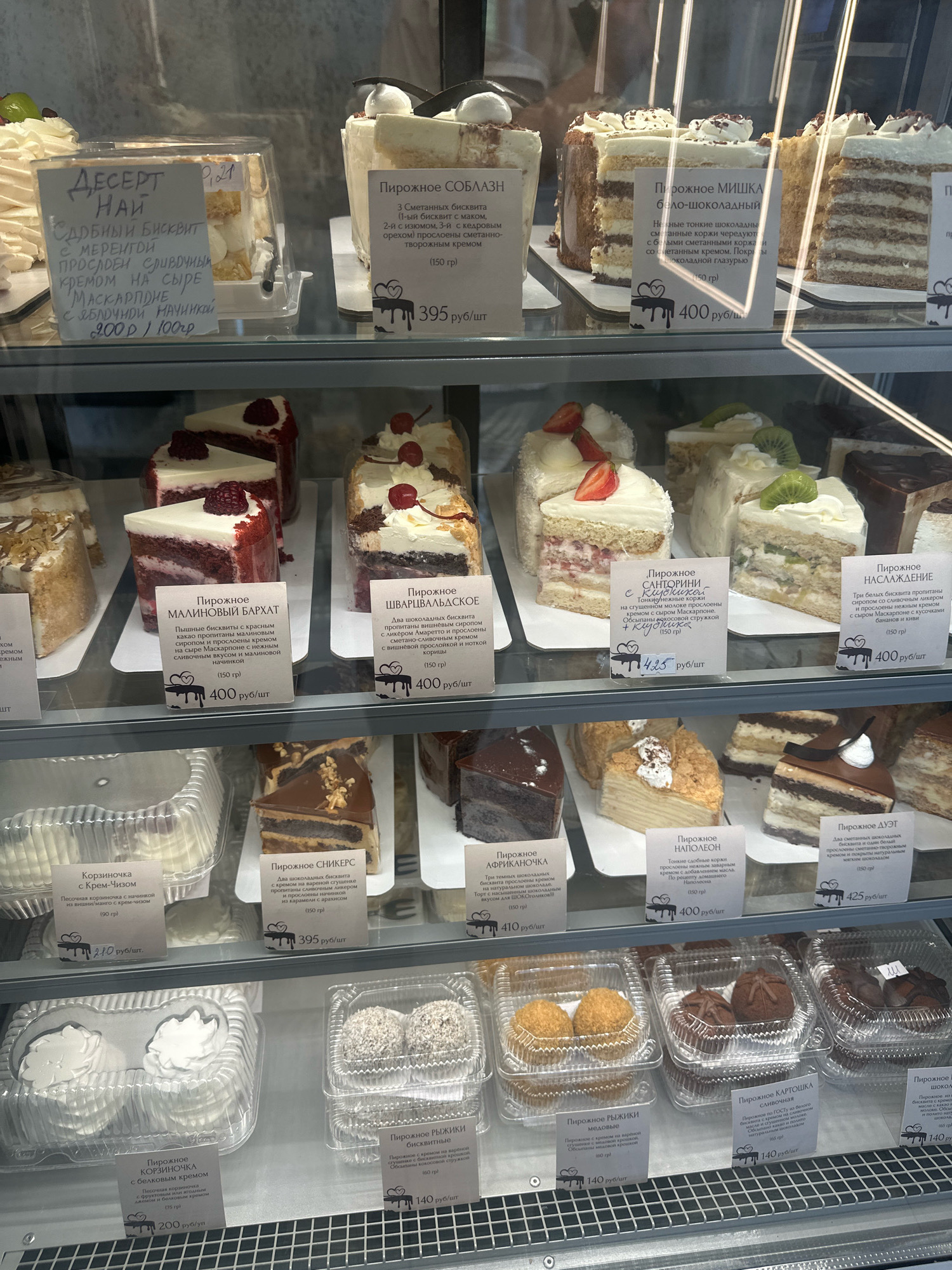 The image shows a display case filled with various cakes and pastries in a confectionery shop. The cakes are arranged on multiple shelves and presented in clear plastic containers or on individual plates. Each item has a small white card with its name and price written in Russian. The shop appears to be well-lit, highlighting the vibrant colors and textures of the desserts. The overall impression is that of a well-stocked and appealing bakery. There are no people visible in the image. The text on the labels indicates the location is Petropavlovsk-Kamchatsky, Russia.