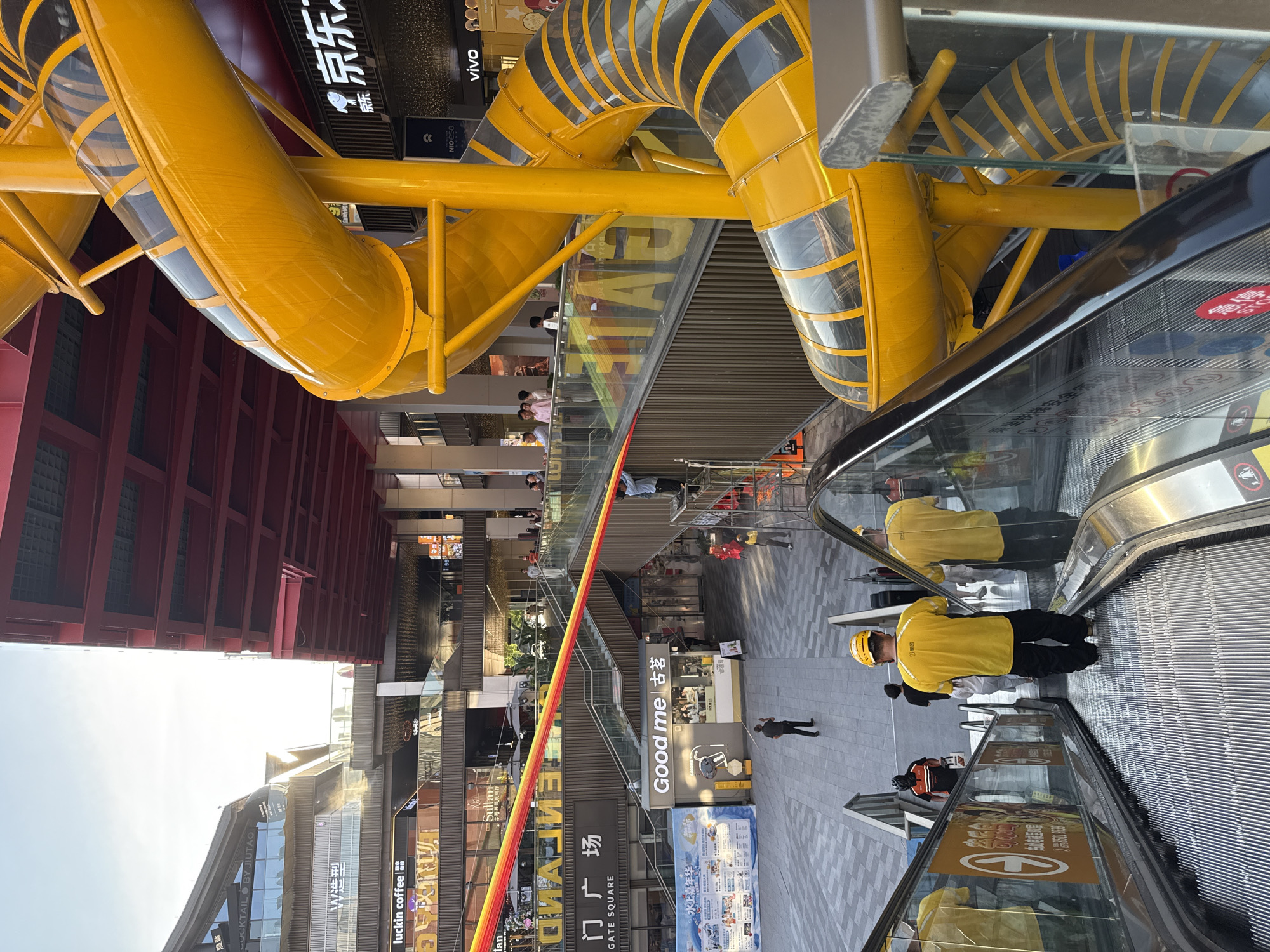 The image depicts an outdoor public space in Yiwu, China, likely a shopping district or plaza, featuring a prominent, bright yellow, spiral slide structure. The slide, constructed of tubular sections with transparent panels, is integrated into a multi-level building complex. In the foreground, an escalator carries several individuals, including at least two wearing yellow shirts and hard hats, suggesting they might be workers. One of these individuals is facing away from the camera, descending the escalator. The background reveals the architecture of the surrounding buildings, which appear to be commercial establishments with signage, including "luckin coffee" and "Good me". A red, elevated structure, possibly a bridge or canopy, is visible overhead. The overall atmosphere suggests a vibrant urban environment during daylight hours.
