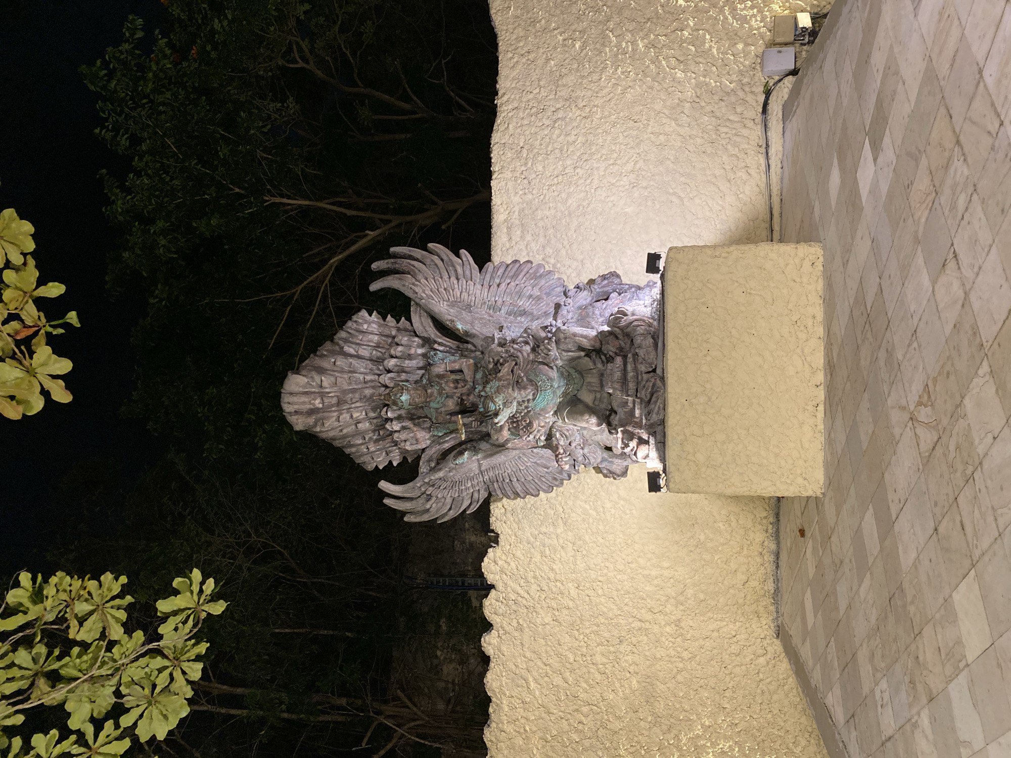 An ornate stone statue of a Garuda bird with a human-like figure seated upon it is centrally located in an outdoor tiled courtyard at night. The statue is illuminated by spotlights, casting a glow on its detailed carvings and green patina. Behind the statue, a textured, off-white wall stretches across the background, with dark trees and foliage visible above and beyond it. The courtyard floor is paved with light-colored square tiles, arranged in a diamond pattern. In the foreground, the edges of the courtyard and the base of the statue are visible. The overall scene suggests a religious or cultural site, possibly a temple or a significant landmark in Kuta Selatan, Indonesia, given the prominent depiction of Garuda, a mythological bird creature revered in Hindu and Buddhist traditions.