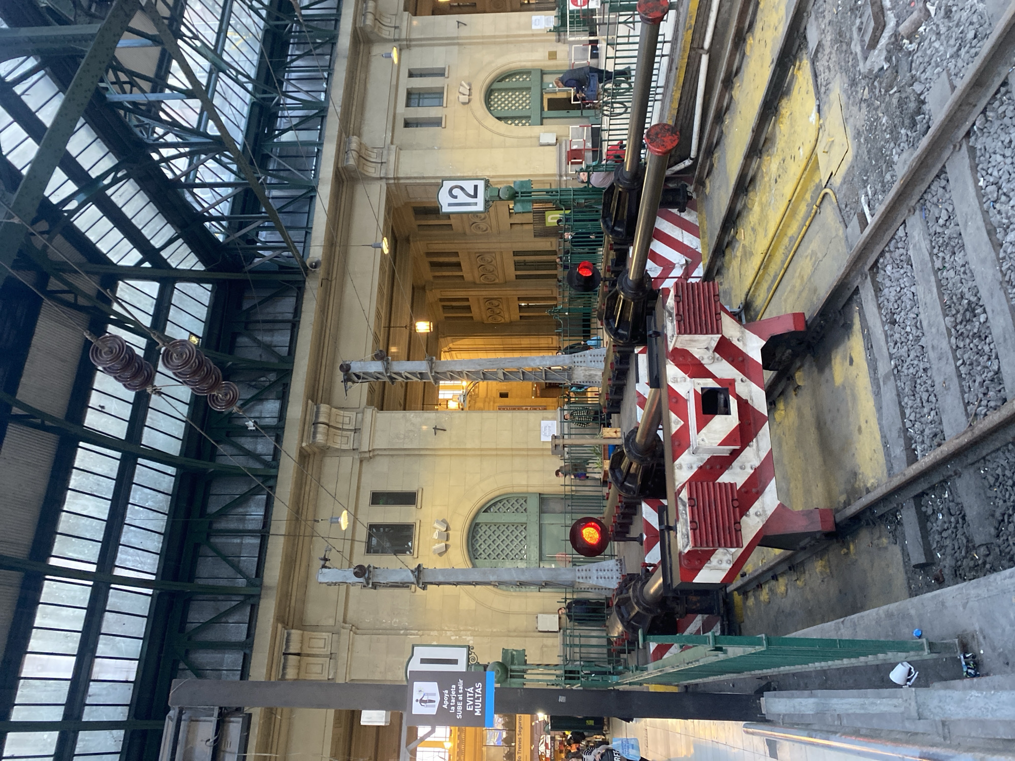 The image shows a train station platform in Buenos Aires, Argentina. Platform 12 is visible in the background. In the foreground are two sets of rail tracks, and adjacent to each track is a striped red and white safety bumper with a signal light illuminated red. A sign in the lower-left portion of the image urges riders to use the SUBE transit card to avoid fines. Overhead, a green metal framework supports the station's glass-paneled roof. The station's architecture features arched windows and detailed molding. There are several people located in the background on the adjacent platform, near platform 12.