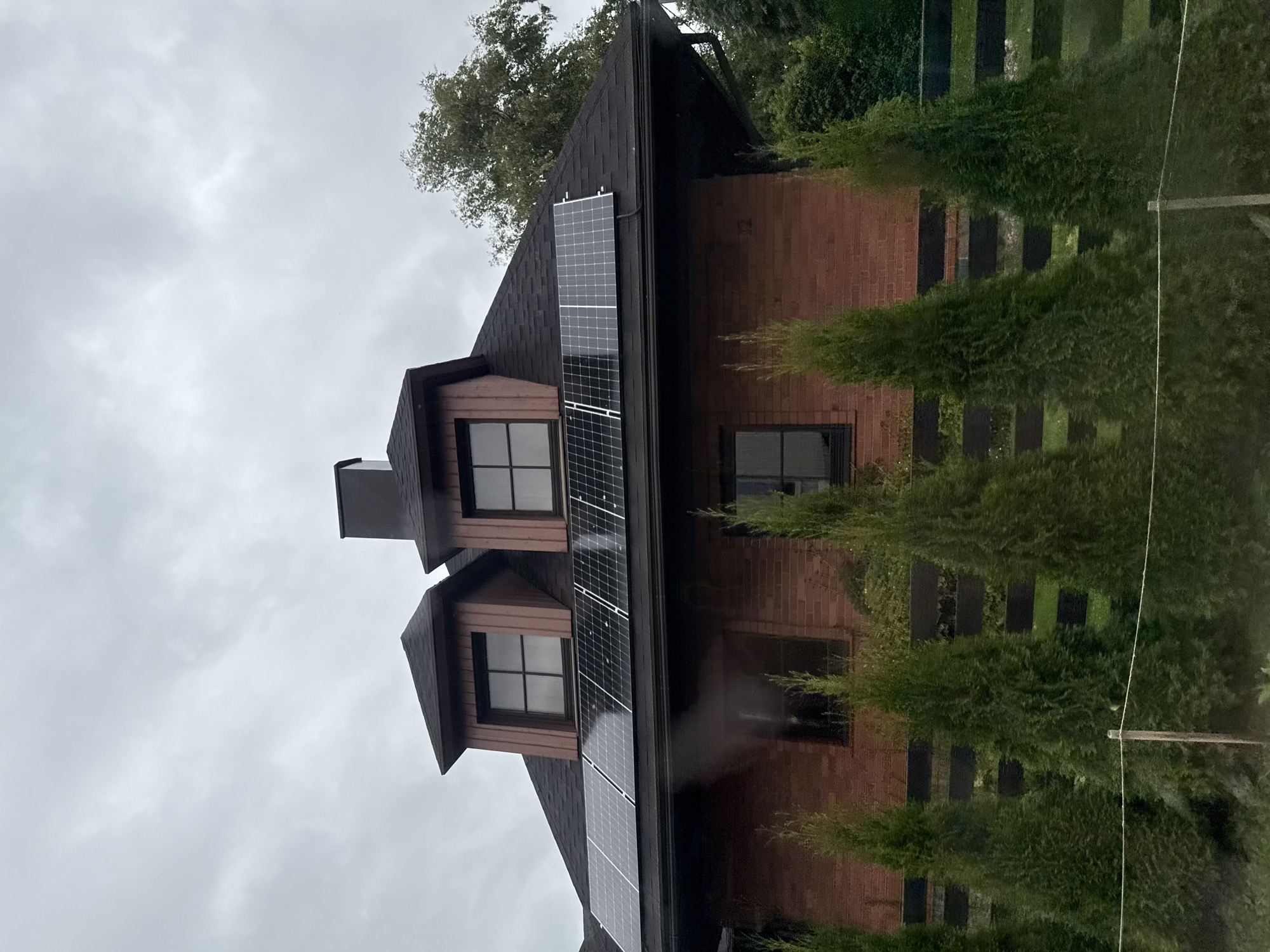 A brick house with a dark roof and dormer windows is shown on a cloudy day. Solar panels are installed on the roof. Green conical trees are planted in front of the house, and a dark fence is visible behind them. The image is an exterior shot and does not contain any people. The location is not specified to be in Tallinn, Estonia.