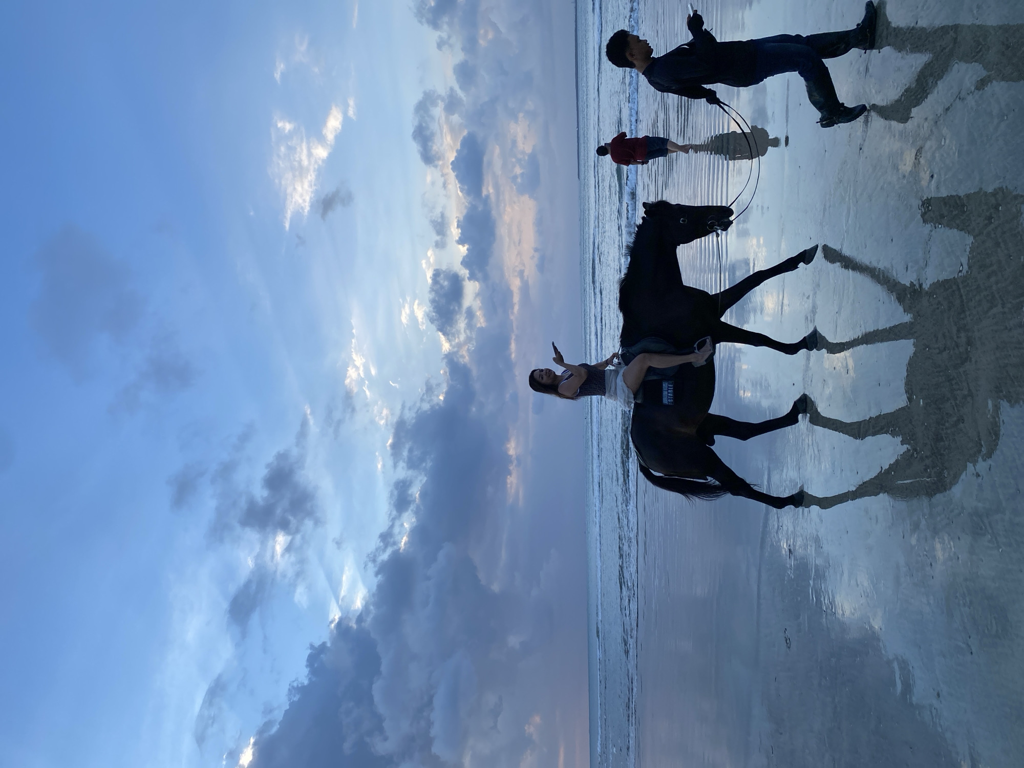 The image captures a serene beach scene in Kuta Selatan, Indonesia, during sunset or dusk. The sky is a prominent feature, filled with dramatic clouds ranging from soft blue to darker grey, with patches illuminated by warm, golden light, hinting at the sun setting. This light creates subtle rays visible through the clouds.
In the foreground, a person, likely a woman, is seen riding a dark-colored horse (possibly black or dark brown) on the wet sand. She wears a light top and appears to be smiling, looking towards the left side of the frame. The horse is equipped with a saddle, on which the text "HUMAN MADE STABLE" is visible, and a bridle. Another person, a man dressed in dark clothing, stands next to the horse, holding its lead rope and appears to be holding a smartphone.
Further out in the shallow ocean water, two more individuals are present. One man in dark attire stands facing away from the camera, while another in a red top is bending over. Gentle waves break along the shoreline. The wet sand acts as a mirror, creating strong, clear reflections of the horse, the people, and the vibrant sky, enhancing the depth and beauty of the scene. The overall mood is peaceful, capturing an evening of horseback riding and enjoyment by the sea.