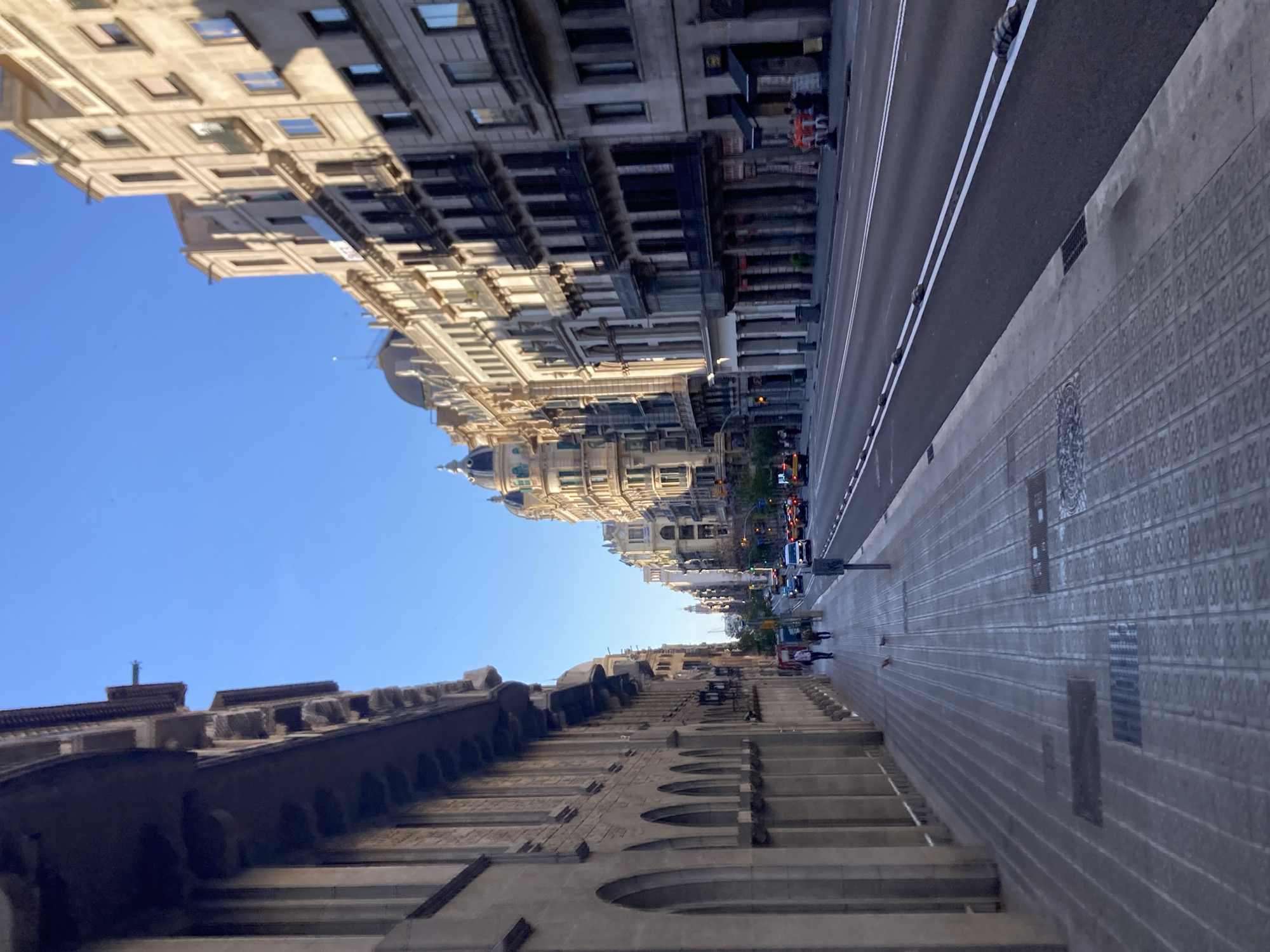 The image captures a view of a wide, paved street in Barcelona, Spain, lined with ornate, multi-story buildings on both sides. The perspective is from a sidewalk on the left, looking down the street towards a distant intersection. The sky is clear and blue.
The building on the left features a stone facade with arched windows and decorative architectural elements along its upper edge. The sidewalk is made of patterned paving stones and has manhole covers and grates. Several people are walking on the sidewalk, some in the mid-ground and others further away.
The street is paved with asphalt and marked with double white lines. Several cars and taxis are visible on the road, moving in the same direction as the viewer. The buildings on the right showcase a variety of architectural styles, with balconies, numerous windows, and intricate detailing. Some of these buildings have rounded domes or turrets.
In the distance, the street curves slightly, and more buildings are visible. There are some streetlights and traffic signals. On the right side of the street, near the foreground, there are a few small tables and chairs set up outside what appears to be a business. The overall impression is of a vibrant, urban street scene in Barcelona during daylight.