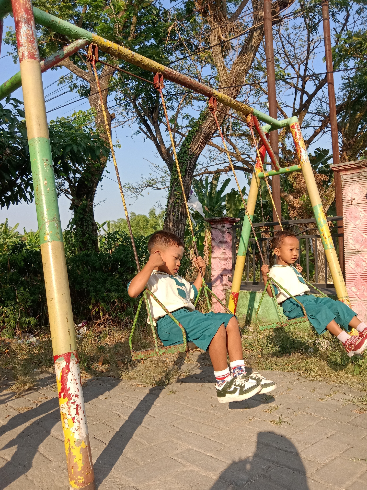 Two young children, appearing to be of Asian ethnicity, are seated on swings of a rusty playground structure. Both children are wearing school uniforms consisting of white shirts with green trim and green shorts or skirts. The child on the left swing is facing to the right and is leaning back, with their left hand resting on their chin. The child on the right swing is facing forward and looking to the left. The swings are suspended by yellow ropes from the metal frame, which is painted with red, yellow, and green colors, exhibiting signs of wear and tear. The playground is situated outdoors on a paved area with some grass and dirt. In the background, lush green trees and foliage are visible under a clear blue sky. The scene appears to be set in a residential or school area in Sugihwaras, Indonesia, likely during daylight hours as indicated by the sunlight casting shadows on the ground.