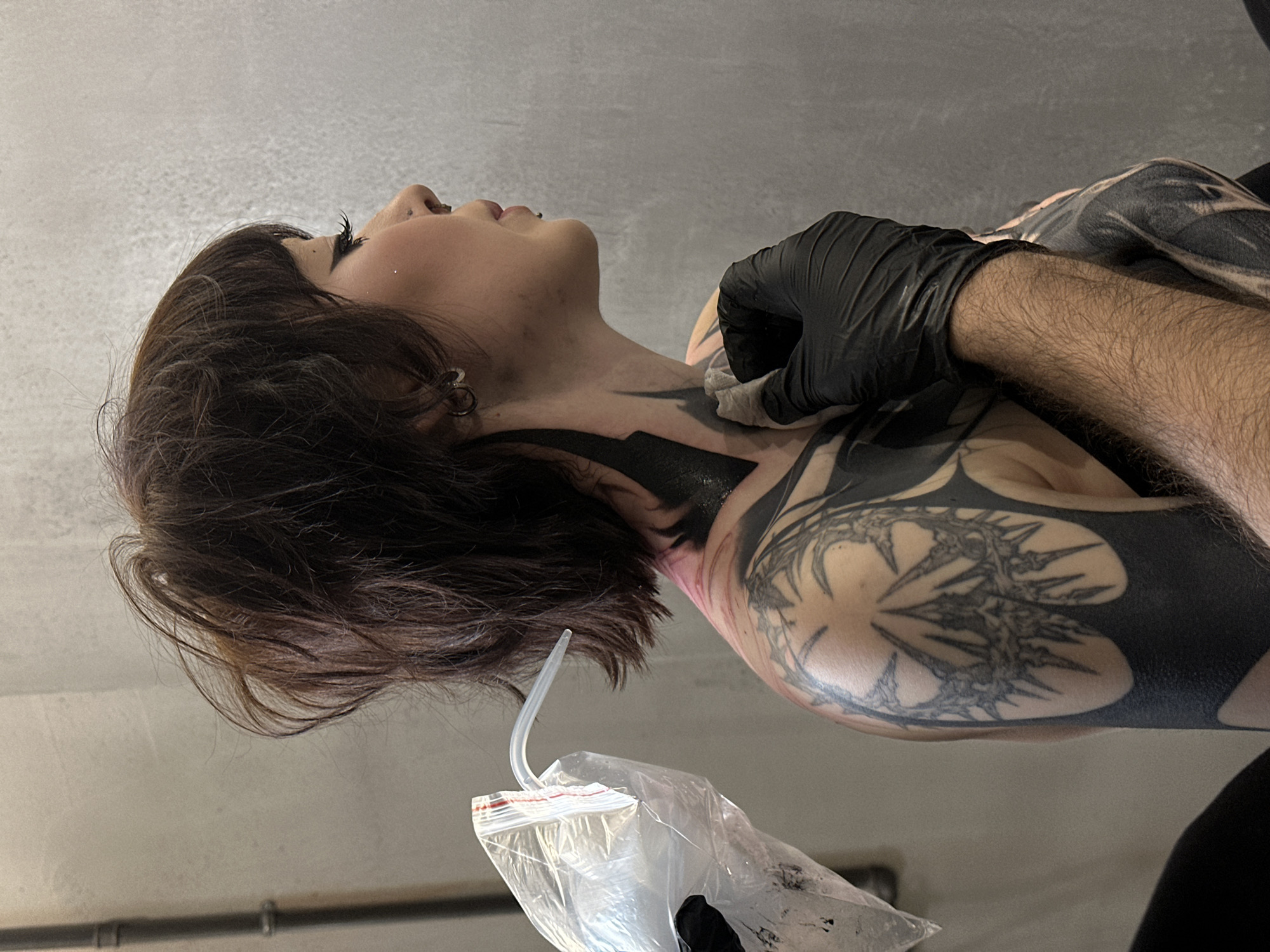 A person lies on their back with their head turned to the left, receiving a tattoo on their neck and upper chest. The person has short, dark brown hair with bangs, and their skin is light-toned. They have a piercing in their ear. A tattoo artist, wearing a black glove, is cleaning the freshly inked area on the person's neck with a white cloth. The tattoo consists of thick, black designs with some areas filled in solid black and others featuring detailed, geometric patterns. The artist's arm is hairy and also covered in tattoos. A plastic bag with a curved tube sticking out of it is visible near the bottom left corner of the image. The background shows a plain, light gray wall with a horizontal metal pipe running along it. There is no explicit evidence to suggest that the location is in the city of Moscow, Russia.