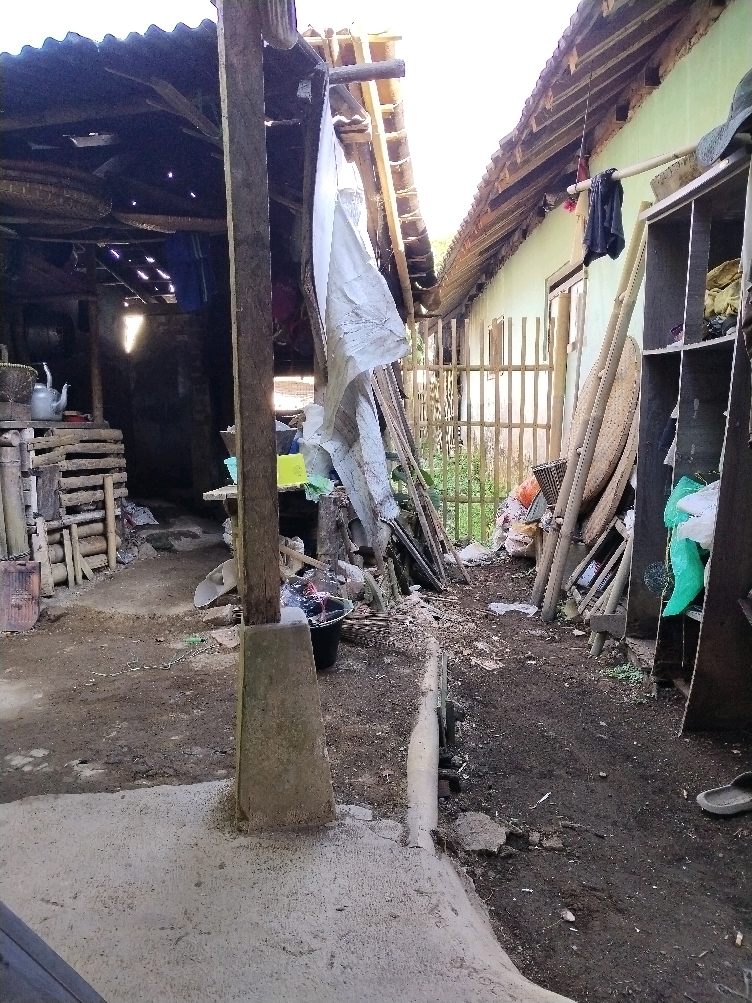 The image depicts an outdoor scene in Desa Cidugalen, Indonesia, showing a cluttered, open-air space between two structures. On the left, a dark, enclosed area with a corrugated metal roof is visible, supported by wooden beams. Various items, including baskets and what appears to be a metal kettle, are placed on a wooden platform. A large, rough concrete post stands in the foreground, obscuring part of the view into the left structure.
To the right, a long, light green building with a tiled roof extends into the background. In front of this building, a tall, narrow structure with shelves, possibly a cabinet or storage unit, is laden with items, including green plastic bags and fabric. Leaning against this structure and the building are several long poles and large, flat circular objects, likely bamboo or woven materials. A wooden fence runs along the base of the green building.
The ground is predominantly dirt and debris, with scattered pieces of wood, plastic, and other refuse. A black bucket, a broom, and a single grey sandal are also visible on the ground. The overall impression is one of a utilitarian and somewhat disorganized environment, likely a residential or working outdoor area. The lighting suggests it is daytime, with some areas in shadow and others illuminated by sunlight.