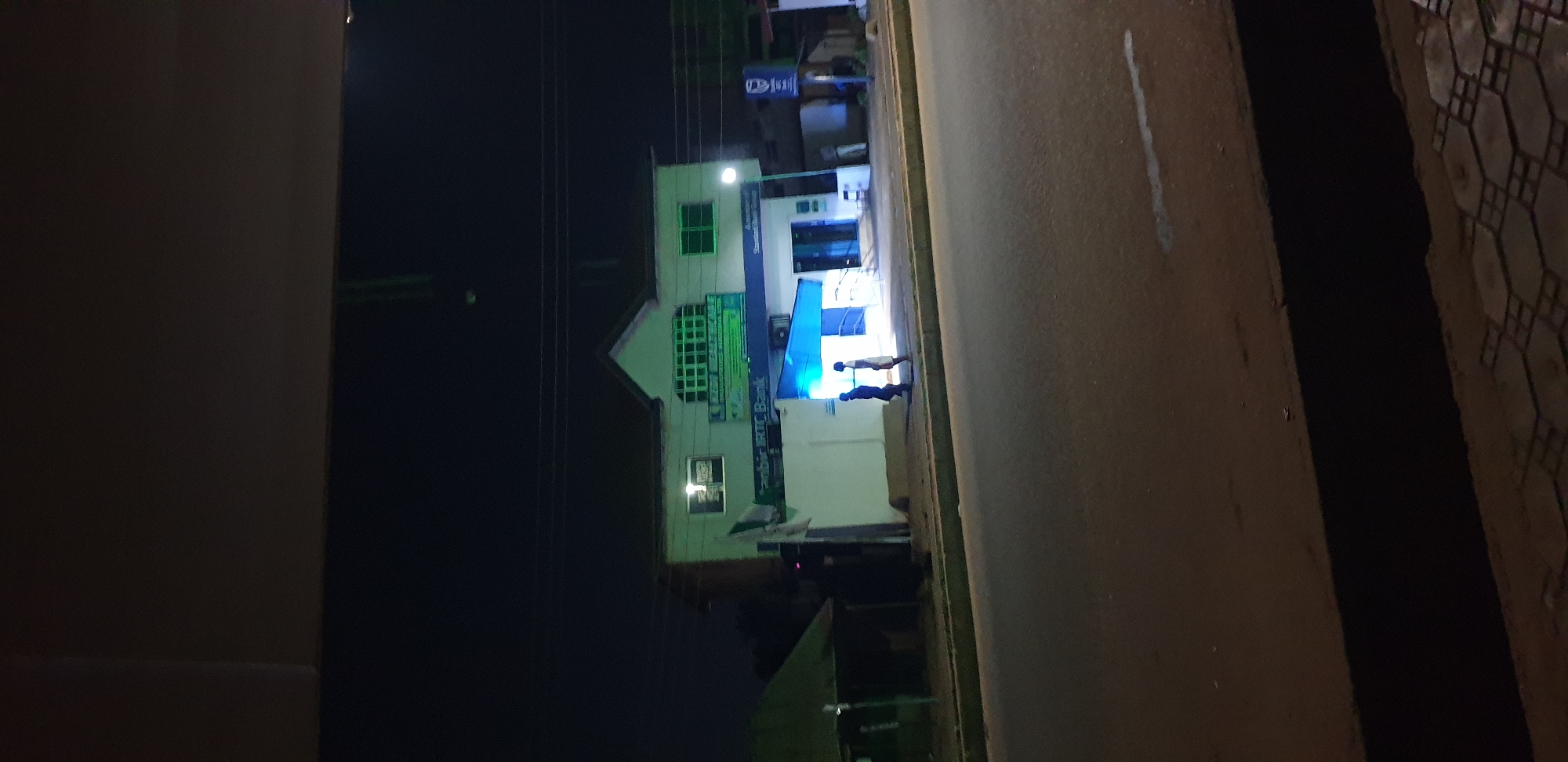 The image captures a nighttime scene, looking across a road towards a commercial building. The perspective is rotated 90 degrees counter-clockwise.
The primary subject is a multi-story building, likely a bank or business, illuminated by various artificial lights. The building is light-colored, featuring several windows, some of which appear to have a green tint or reflection. A prominent green sign with white text is visible on the upper part of the building, though the text is largely unreadable due to blur and angle. Below this, a long blue sign runs horizontally across the building facade. Portions of this sign read "Sabr for Bank" and possibly "Standard Chartered Bank," given the recognizable blue branding and style. A smaller blue sign with a logo resembling "DBS" or a similar banking institution is visible higher up on the building.
Below the main signs, near what appears to be an entrance, two people are standing. They are silhouetted against a bright blue light emanating from beneath an awning or structure at the building's entrance. One person is taller than the other. Their activity is unclear, but they appear to be either waiting, entering, or exiting the building.
To the right of the building, a paved road or wide pathway stretches into the distance, with a faint white line marking part of its surface. Further to the right, a textured ground surface, possibly patterned paving stones or tiles, is visible. The sky is dark, indicating it is nighttime. Overhead, several power lines run horizontally across the frame. The overall lighting is dominated by the artificial illumination from the building and the adjacent street. The scene suggests a commercial area after dark in Sabon Tasha, Nigeria.