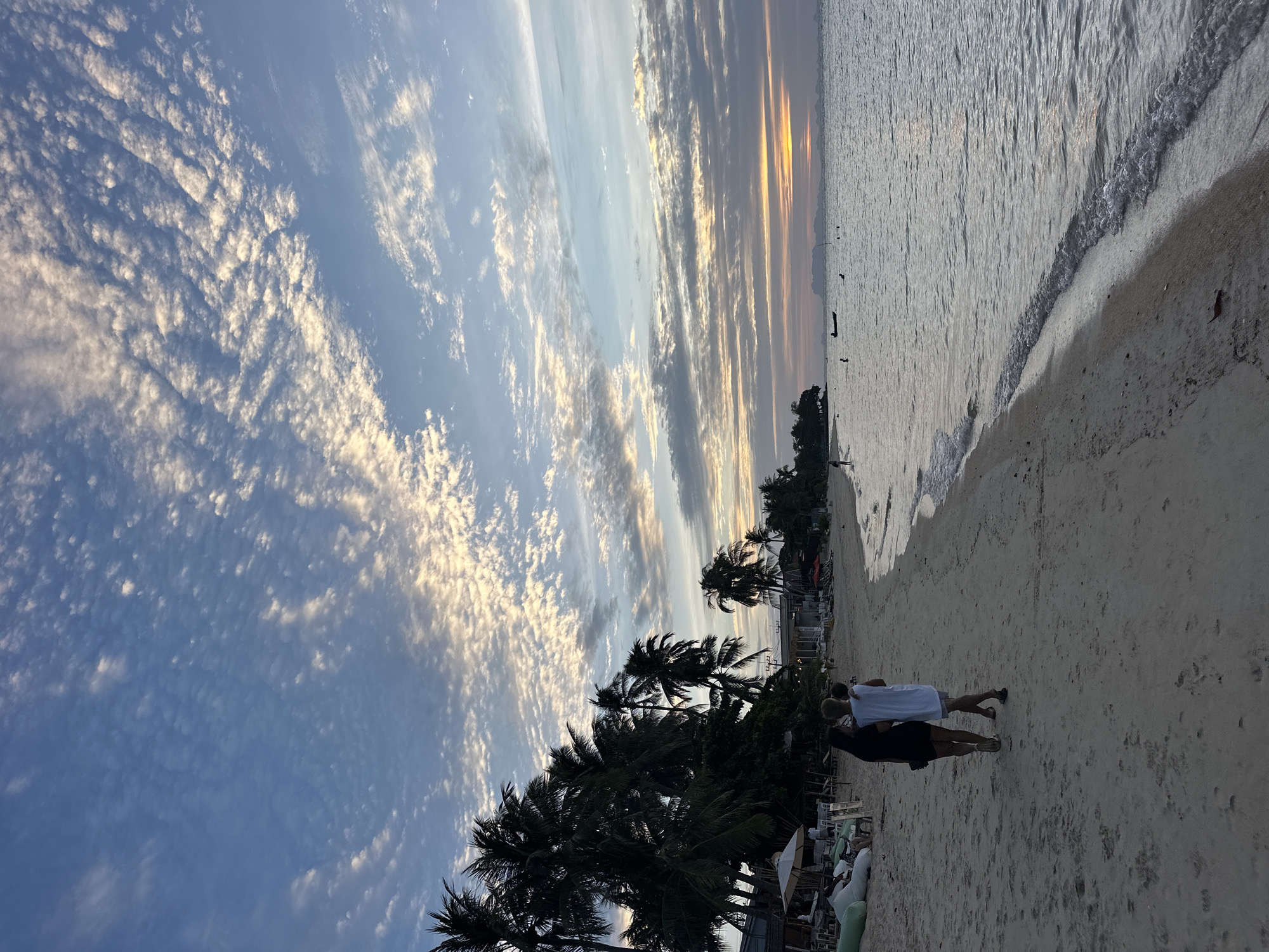 A family walks along the beach in Ko Samui District, Thailand, during a beautiful sunset. The sky is a canvas of soft blues and whites, with scattered clouds tinged with the warm hues of the setting sun. The ocean is calm, with gentle waves lapping at the shore. Palm trees line the edge of the beach, their fronds silhouetted against the sky. In the distance, buildings and other beachgoers suggest a vibrant coastal community. The family, a mother and two children, walk hand-in-hand, enjoying the serene evening. The scene is peaceful and idyllic, capturing the essence of a tropical paradise.