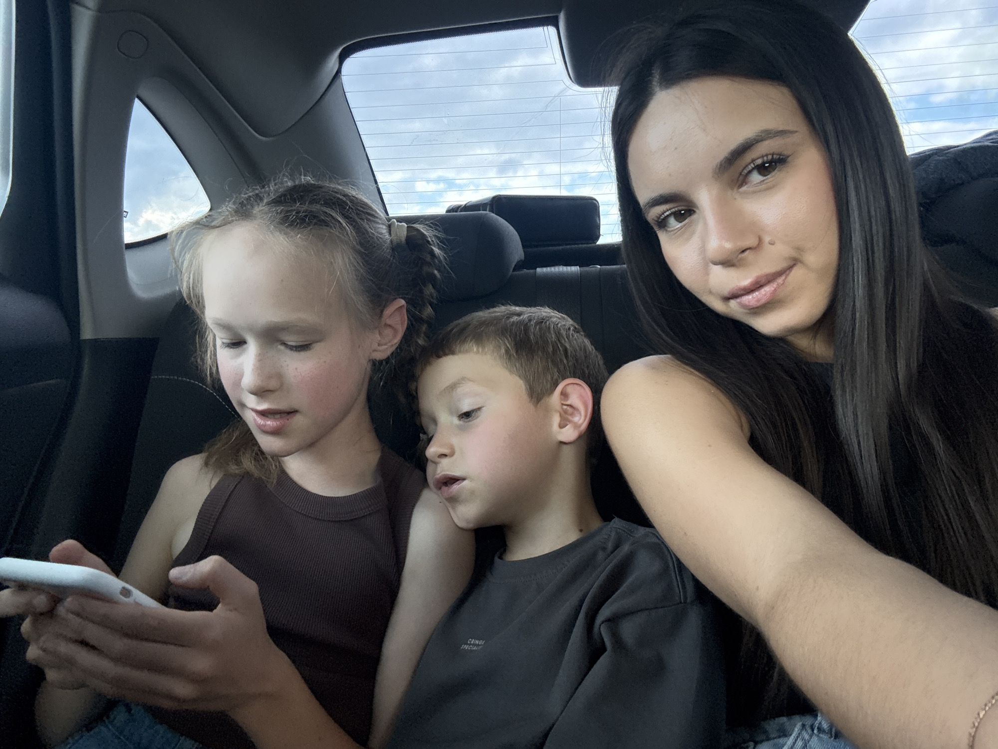 In the image, three individuals are seated in the back of a vehicle, as indicated by the car's interior upholstery and windows visible. A young woman with long, dark hair is in the foreground on the right, looking towards the camera. To her left, a young girl with braided blonde hair is looking down at a smartphone she is holding. Next to the girl, a young boy with brown hair is also looking down. The interior of the car is dark, suggesting it is daytime. The background shows a cloudy sky through the rear window. The scene appears to be a casual moment captured inside a car.