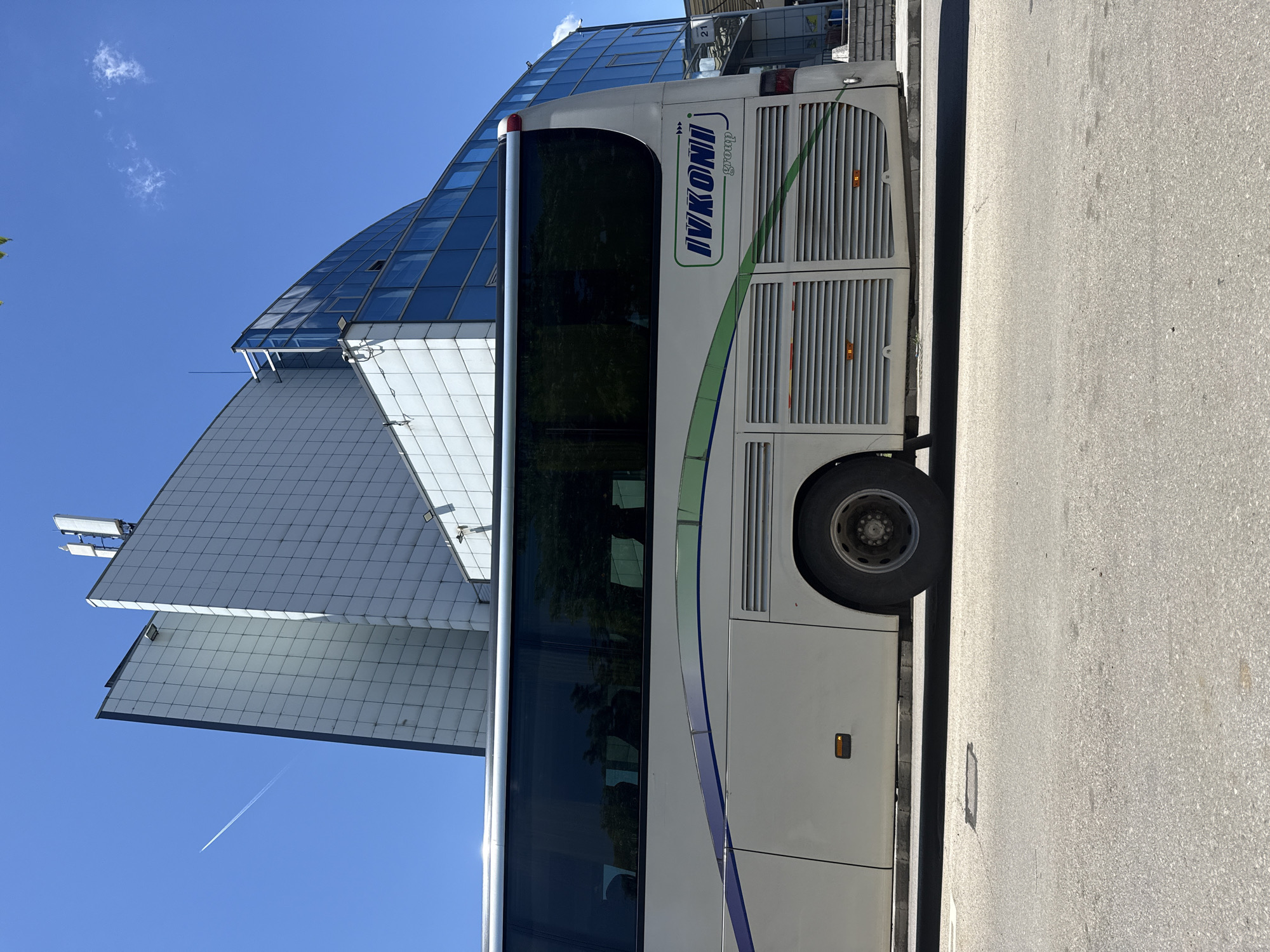A white bus with "IVKONI group" branding is parked on an asphalt road in front of a modern building with a glass facade. The building has multiple sections, some with a tiled exterior and others with large blue glass panels. The sky is clear blue with a faint contrail from an airplane visible. The bus has tinted windows and green and purple decorative stripes along its side. A portion of the bus's wheel and fender are visible, as well as ventilation grates on its rear section. The image appears to be taken during daylight.