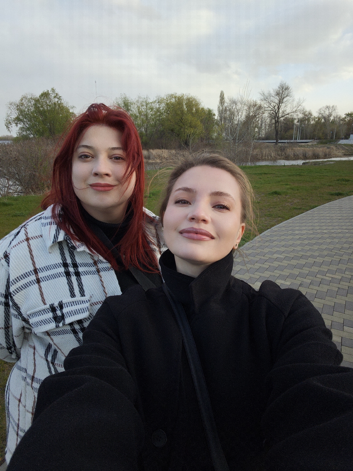 An outdoor selfie features two young women smiling into the camera, set against a natural landscape in Kyiv, Ukraine. The woman on the left, with vibrant red hair, is positioned slightly behind and to the side, wearing a white and black plaid shacket over a black top. She looks towards the camera with a gentle, pleasant smile. The woman taking the photo, on the right, has light brown hair swept back and is dressed in a black overcoat, her expression relaxed and happy as she gazes into the lens.
The background shows a serene outdoor environment, likely a park or riverside. A mix of trees, some with emerging green leaves and others still bare, suggests the season is late winter or early spring. A narrow body of water, possibly a river or canal, runs through the mid-ground, bordered by green grass and reeds. A distinctive paved pathway, made of alternating grey and beige rectangular blocks, curves away into the distance on the right side of the image. The sky above is cloudy and overcast, providing soft, diffused lighting, indicative of an afternoon or early evening. The overall mood is casual and friendly. No text is visible within the image.