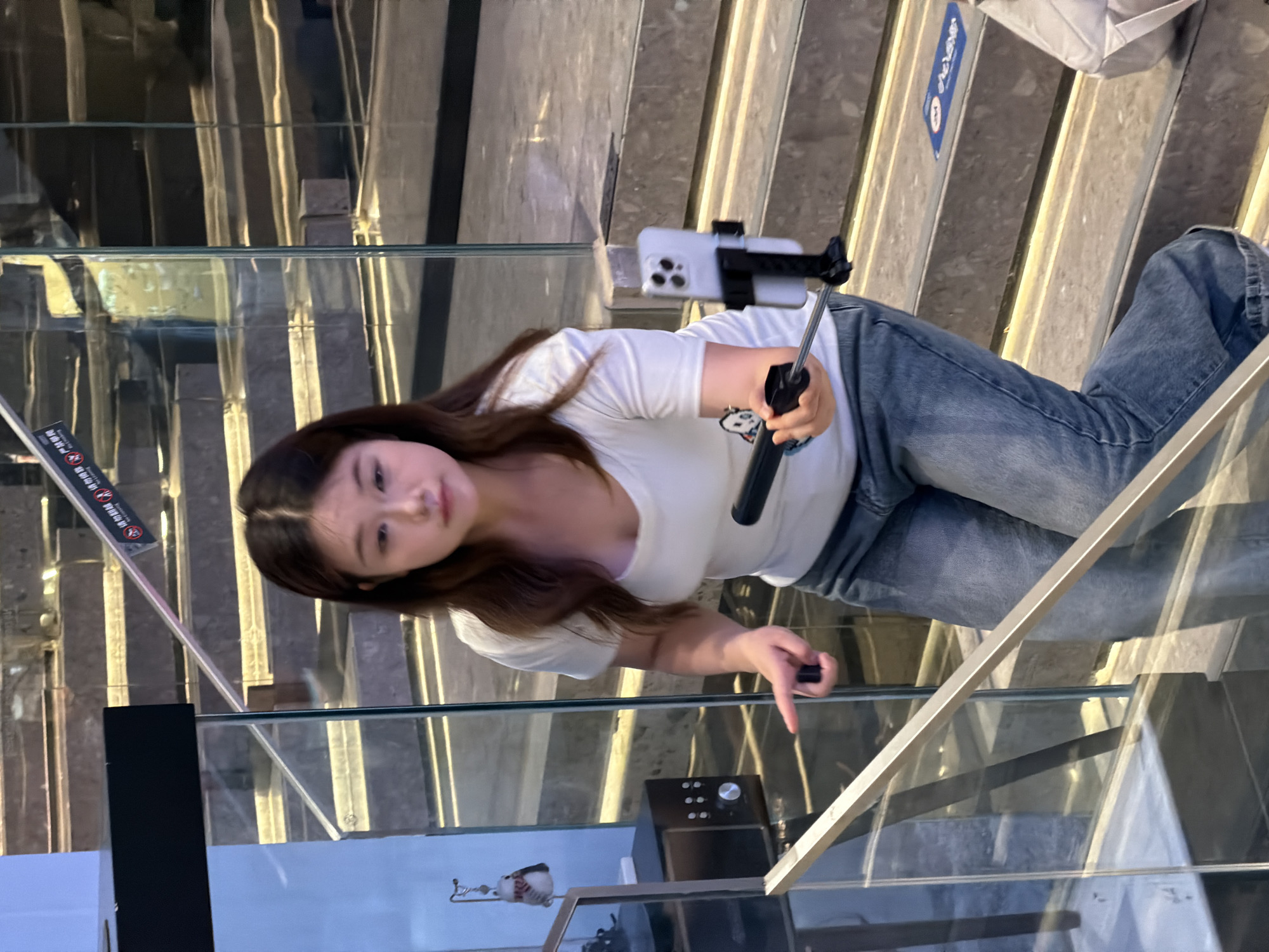 A young East Asian woman is standing on a marble staircase with illuminated risers, holding a selfie stick with a smartphone attached. She is wearing a white V-neck t-shirt and blue denim jeans. Her right hand is extended, seemingly interacting with the selfie stick. The background shows more of the staircase, with glass railings and some signage on the steps. The scene appears to be an indoor public space, possibly a shopping mall or a hotel in Yuzhong District, China, given the context provided.