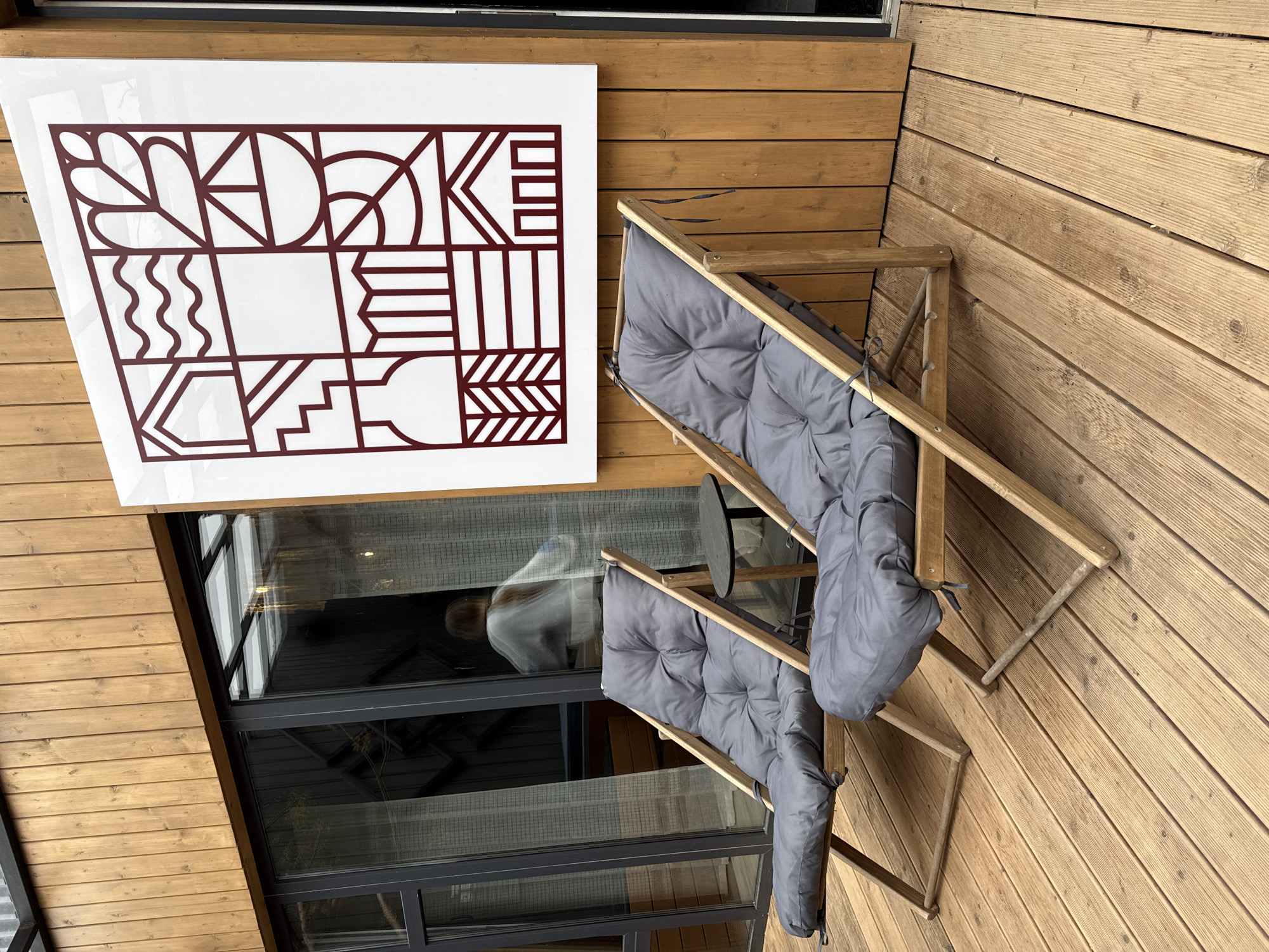 The image shows an outdoor scene, likely a balcony or patio, featuring wooden decking. Two folding lounge chairs with gray cushions are positioned on the decking. A white square sign with geometric maroon-colored designs is mounted on the adjacent wall. The wall also contains a large window with dark framing, reflecting an interior space with a person visible. The location, Vyakhtelevo, Russia, cannot be confirmed from the image alone.