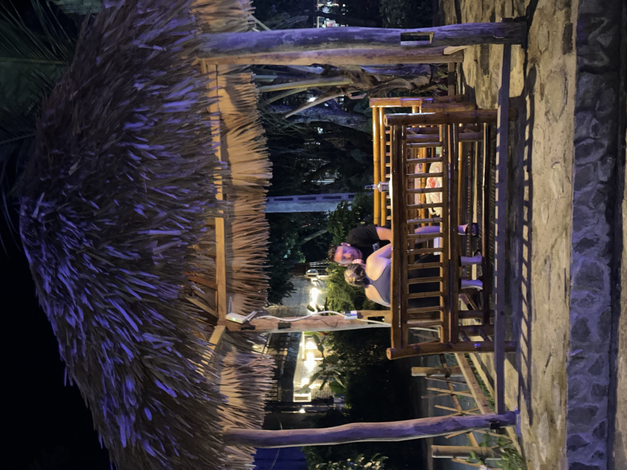 The image depicts an outdoor seating area at night, possibly a restaurant or bar, with a thatched roof supported by wooden poles. In the foreground, a couple is seated on a wooden bench, facing away from the camera. The man, on the right, is wearing a black t-shirt and is looking at the woman. The woman, on the left, is wearing a dark blue tank top and her hair is pulled back. A bottle, possibly of alcohol, is placed on a table or ledge in front of them. The area is illuminated by artificial lights, casting shadows on the stone-like ground. In the background, a building with illuminated windows and lush vegetation can be seen, suggesting a tropical or subtropical environment. The overall atmosphere is relaxed and intimate.