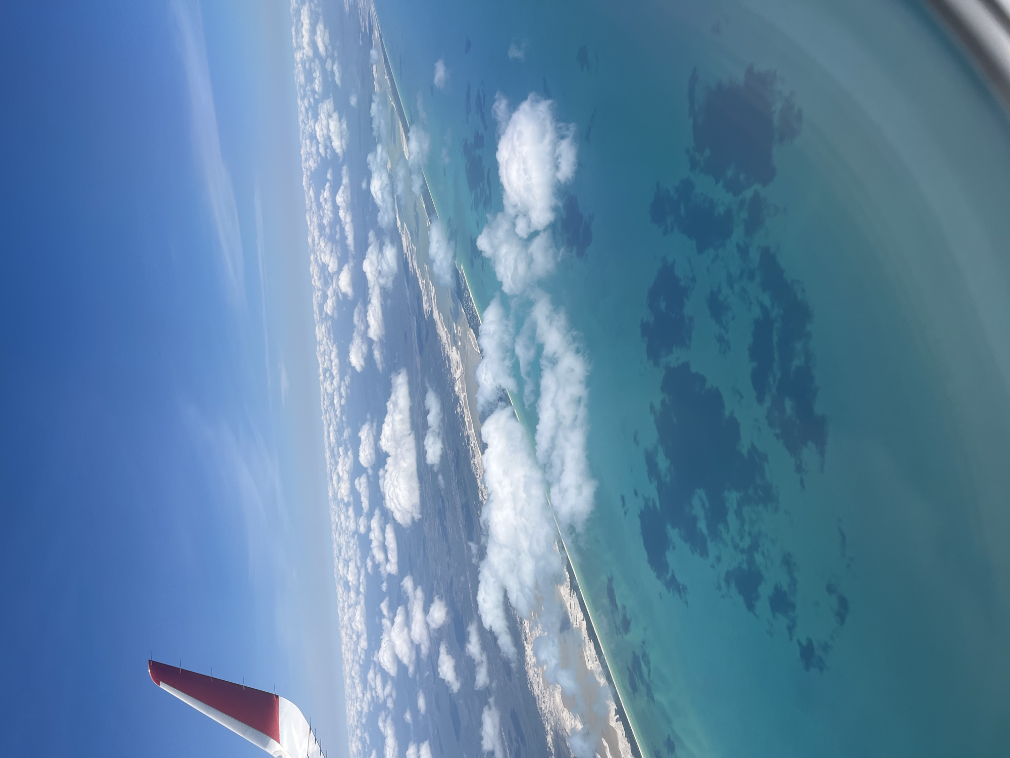 This media file presents an aerial view from an airplane window, capturing a breathtaking tropical coastline under a bright, clear sky. The image is rotated, showing the horizon diagonally, with the deep blue sky occupying the left side and the vibrant turquoise ocean on the right.
In the bottom left corner, a portion of an airplane's wing is visible, painted in white with a red tip and some small antenna-like structures, indicating the vantage point is from a passenger aircraft. Above the wing, the sky is a clear, brilliant blue, with some wispy clouds.
Running vertically through the middle of the frame is a narrow strip of land, a coastline with what appears to be white sandy beaches separating it from the ocean. Scattered over this landmass and extending into the sky are fluffy white cumulus clouds, some casting soft shadows.
The ocean on the right side is a stunning turquoise, characteristic of shallow, clear waters found in tropical regions. Darker patches are visible beneath the water's surface, likely indicating reefs, seagrass beds, or deeper areas. The curved edge of the airplane window is subtly visible in the upper right, creating a slight blur or reflection on that side of the frame.
The bright illumination suggests it is daytime, likely mid-morning or afternoon. The overall scene evokes a sense of serene travel and the natural beauty of coastal landscapes. Given the location context of San Pedro Chenchelá, Mexico, this view is consistent with the Caribbean coast of the Yucatán Peninsula. There is no visible text in the image.