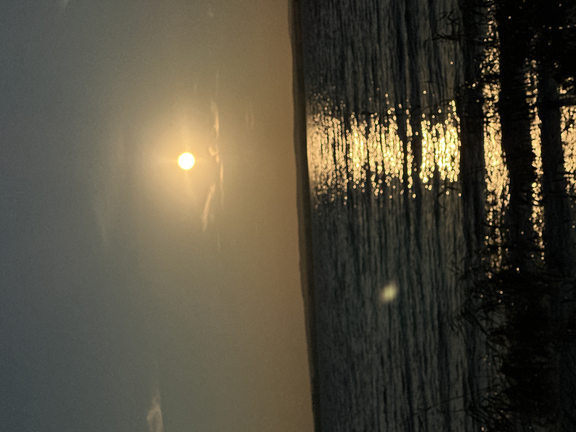 The image depicts a body of water, likely a lake or reservoir, with the sun setting or rising over the horizon. The sun, a bright yellow orb, is partially obscured by a faint haze or clouds in the sky. The light from the sun reflects intensely on the surface of the water, creating a shimmering path of golden light. The water itself appears dark and slightly rippled. In the foreground, silhouetted vegetation, possibly reeds or grass, is visible along the edge of the water. The overall atmosphere is serene and tranquil, characteristic of a natural landscape at dusk or dawn. This scene could be located in Energetik, Russia, a city known for its proximity to Lake Baikal and its surrounding natural beauty.