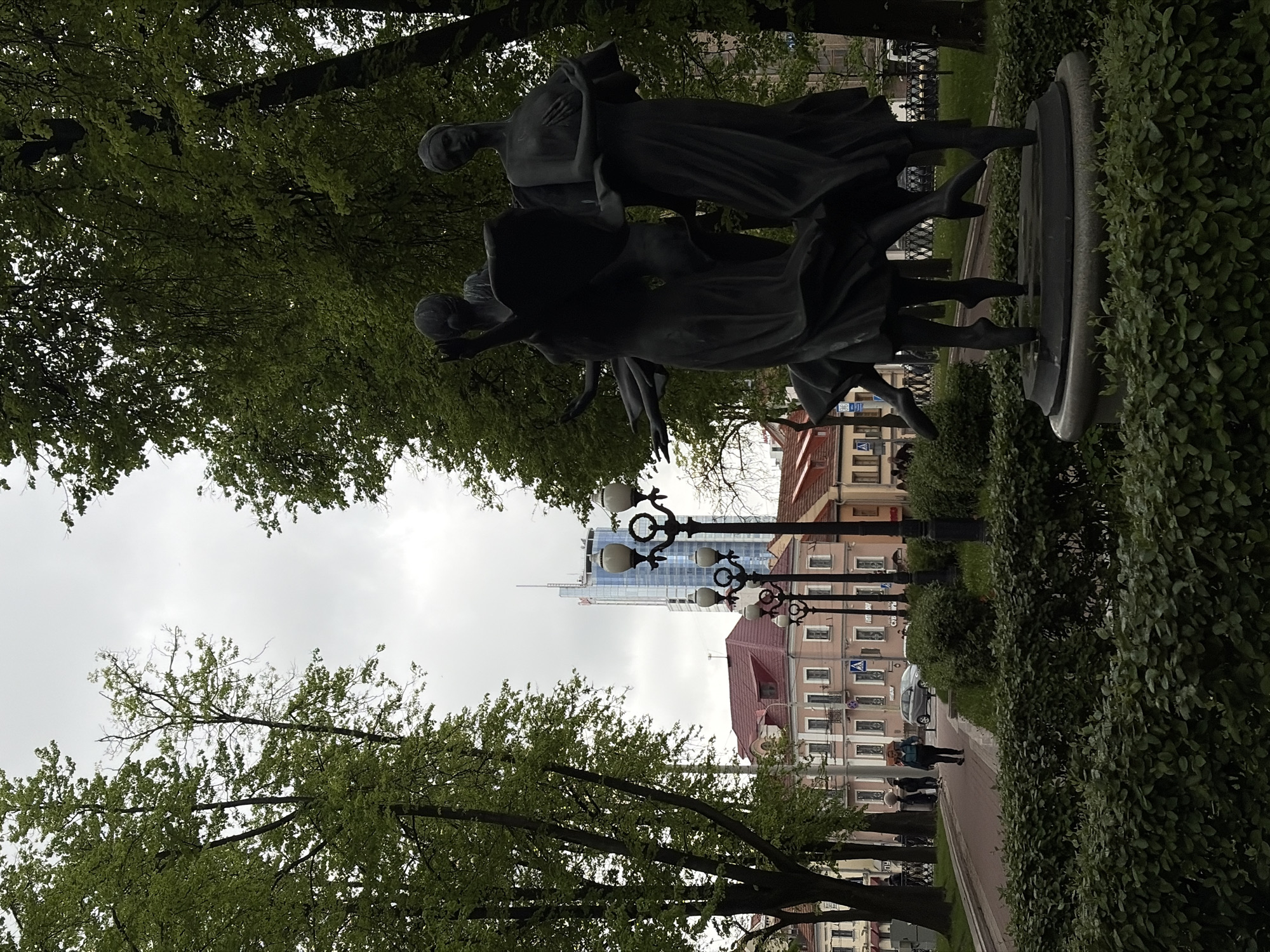 The image shows a park in Unknown, Unknown. A statue of three figures stands on a pedestal amidst green trees and manicured hedges. In the background, a street with light posts lines buildings of varying heights and colors, including a tall modern glass structure. People are walking along the sidewalk. The sky is overcast.
