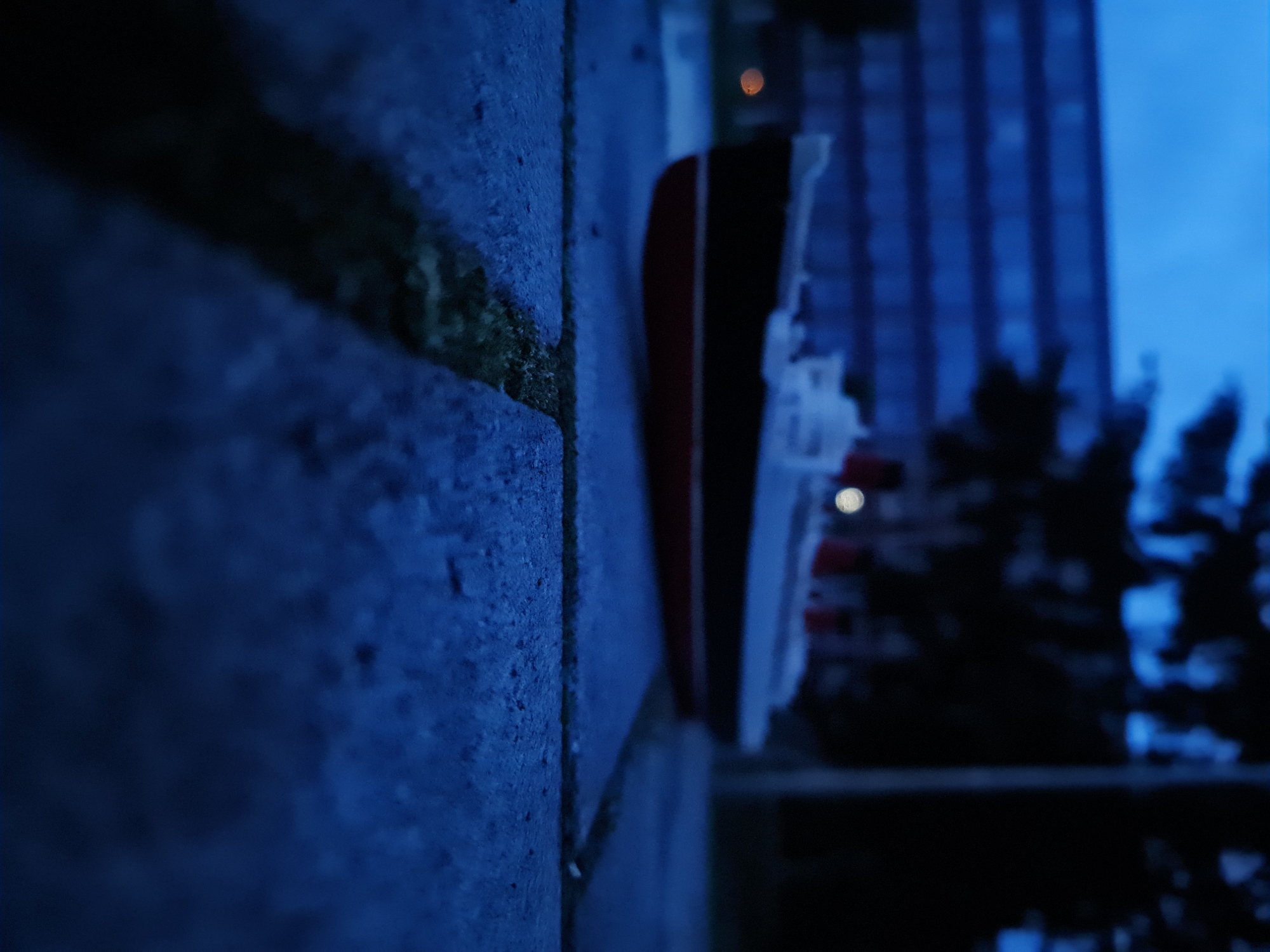 A low-angle, close-up shot features a miniature model of a ship placed on a textured, grey paved surface, possibly a sidewalk or plaza. The ship, predominantly white with a dark hull and a red waterline, is positioned in the mid-ground of the image. In the background, a blurred, multi-story building with horizontal window lines is visible against a dark, overcast sky. The overall lighting suggests either dusk or dawn, with a deep blue hue dominating the scene. The ground in the foreground shows a narrow, dark crevice, suggesting a seam between paving stones, with a hint of green vegetation growing within. The environment is urban and appears quiet, with no people or other vehicles visible. The location is identified as being in the city of Prešov, Slovakia.