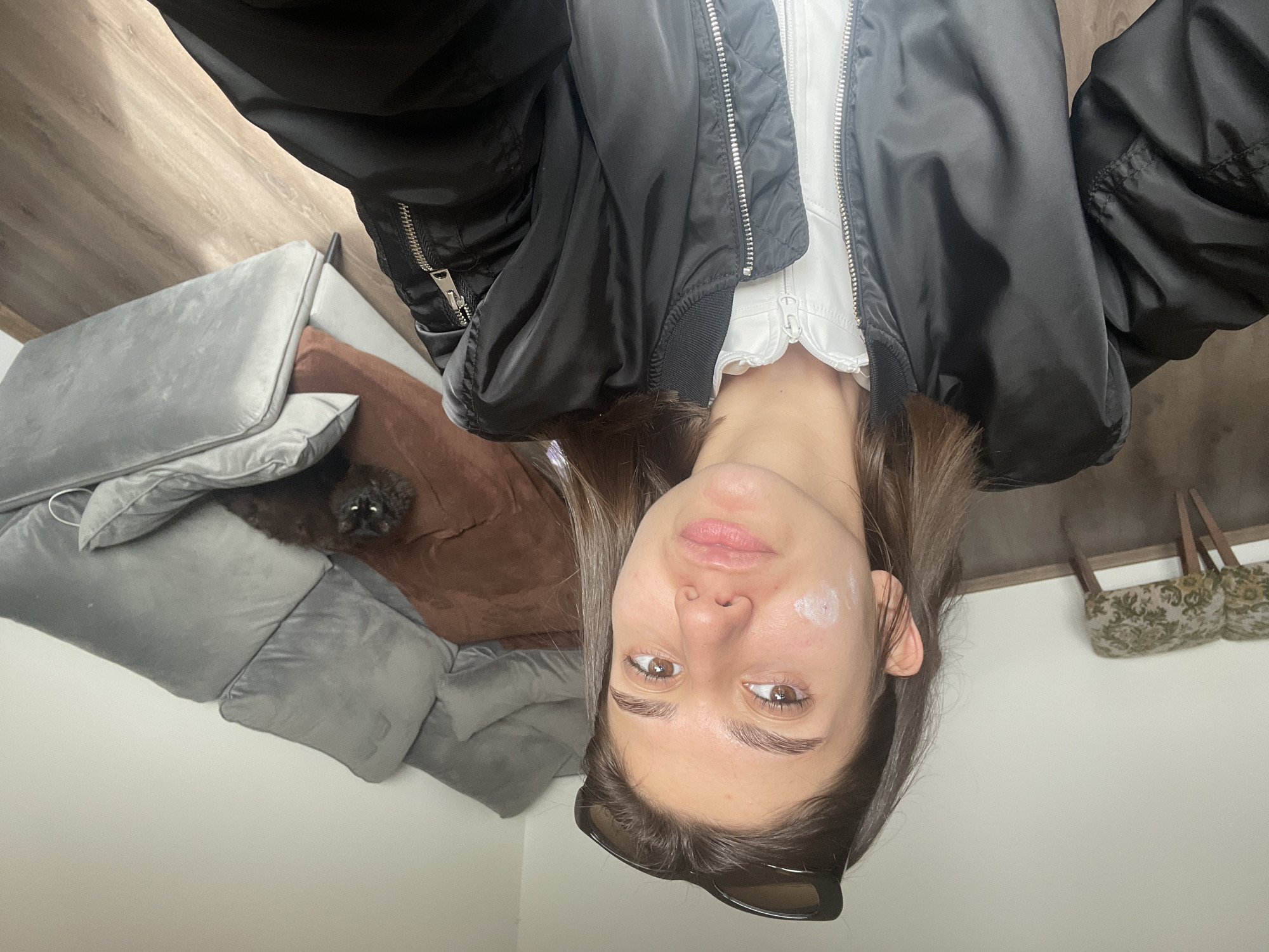 The image, originally oriented upside down, depicts a young woman posing for a photo indoors, with a dog partially visible in the background. The setting is a room, likely in Sofia, Bulgaria.
The young woman is the central subject, looking directly at the camera with a neutral expression. She has medium-length brown hair, and dark sunglasses are resting on top of her head. She is dressed in a black bomber jacket over a white top. A small, circular patch of white cream or product is visible on her left cheek.
In the background, a black dog with bright, reflective eyes is peeking from behind plush grey cushions, which are part of a sofa or bed. A brown cushion or blanket is also present on the furniture. The room features light-colored walls, and a light wooden surface, possibly flooring or wall paneling, is visible. To the right side of the frame, two small, decorative bags with an ornate, light-colored pattern are hanging against the wall. The indoor lighting suggests it could be daytime or illuminated by artificial light. There is no visible text in the image.